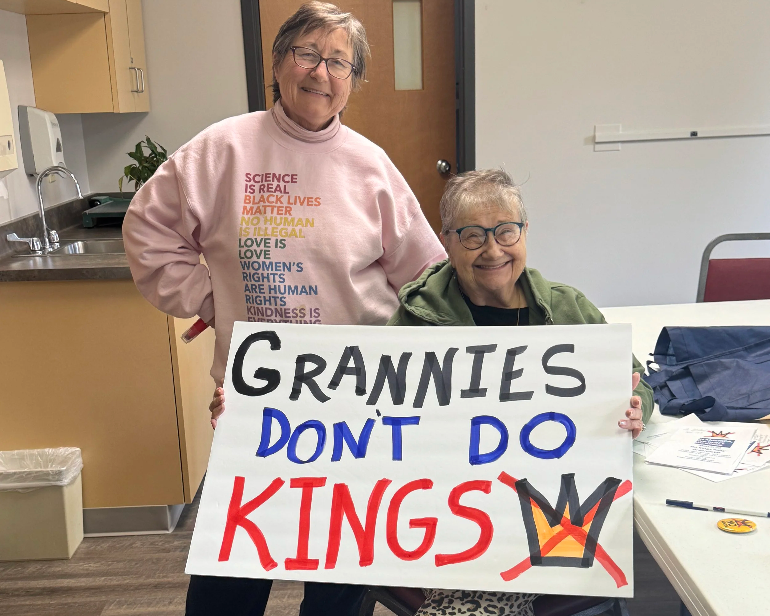 Two women, one elderly and one middle-aged, smiling and holding a sign that says "Grandmies Don't Do Kings" with a crown drawing crossed out. They are in a room with a table, papers, and a counter with a sink in the background.