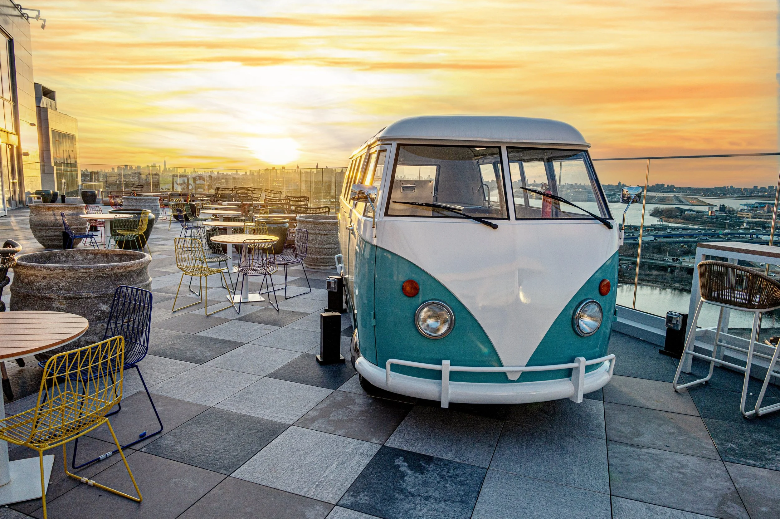 A vintage turquoise and white Volkswagen Type 2 van parked on a rooftop terrace at sunset, with colorful chairs and tables around and a city skyline in the background.