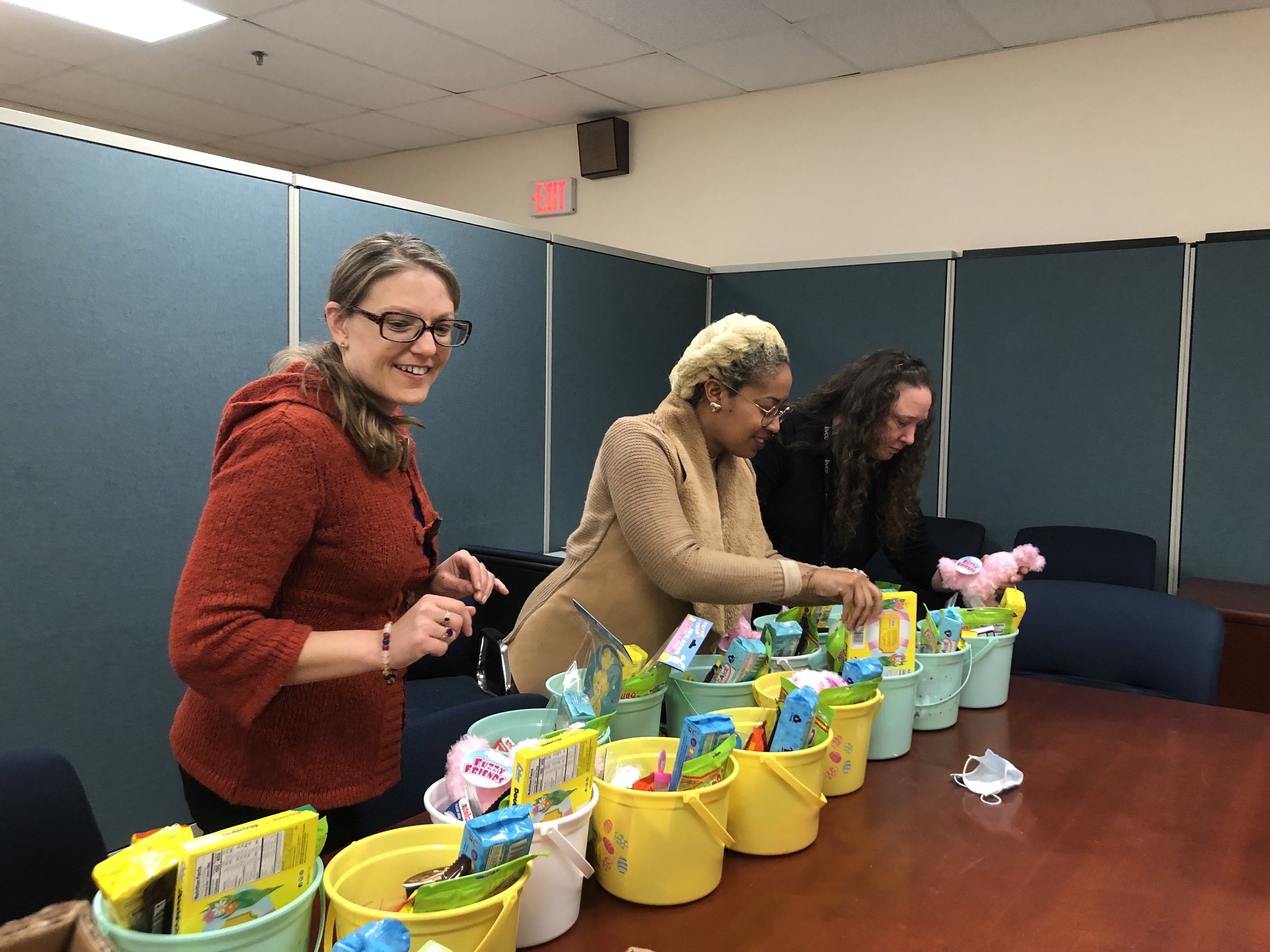Three women standing behind a table filled with colorful Easter baskets and toys, smiling and preparing gifts in a room with blue partition walls.