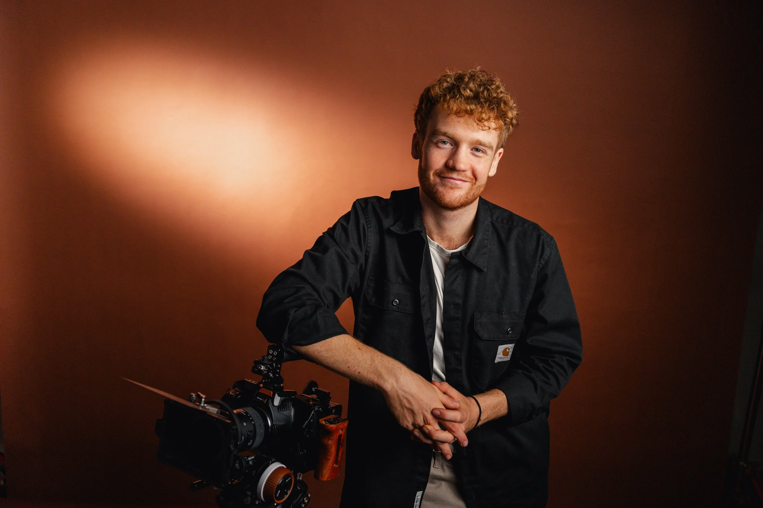 A young man with curly red hair and a beard standing next to a professional camera, smiling, against a gradient brown background.