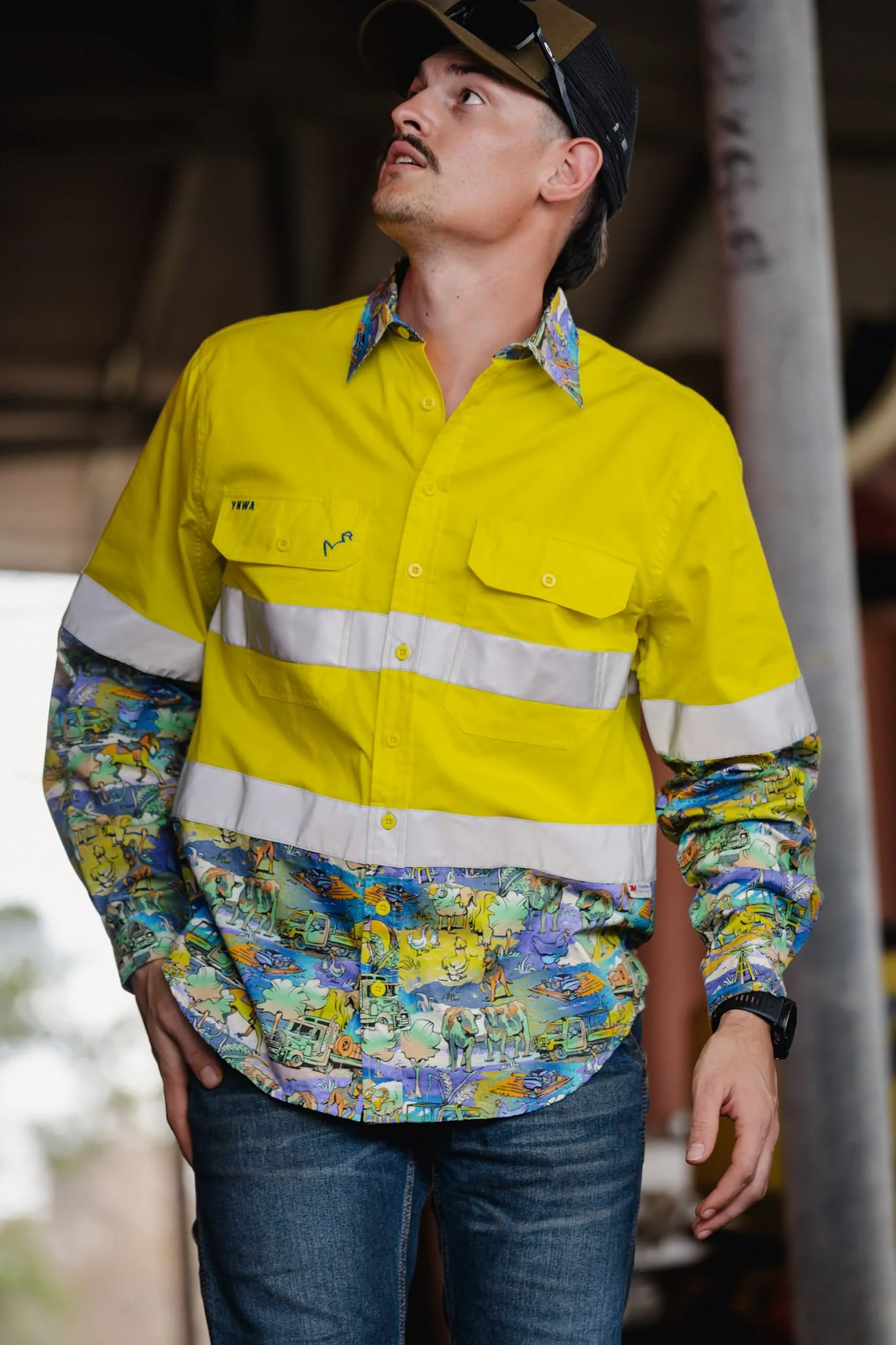 A man wearing a yellow safety vest with reflective strips and a colorful, patterned shirt underneath. He is looking upward and slightly to the side, wearing a black and brown cap and a black watch.