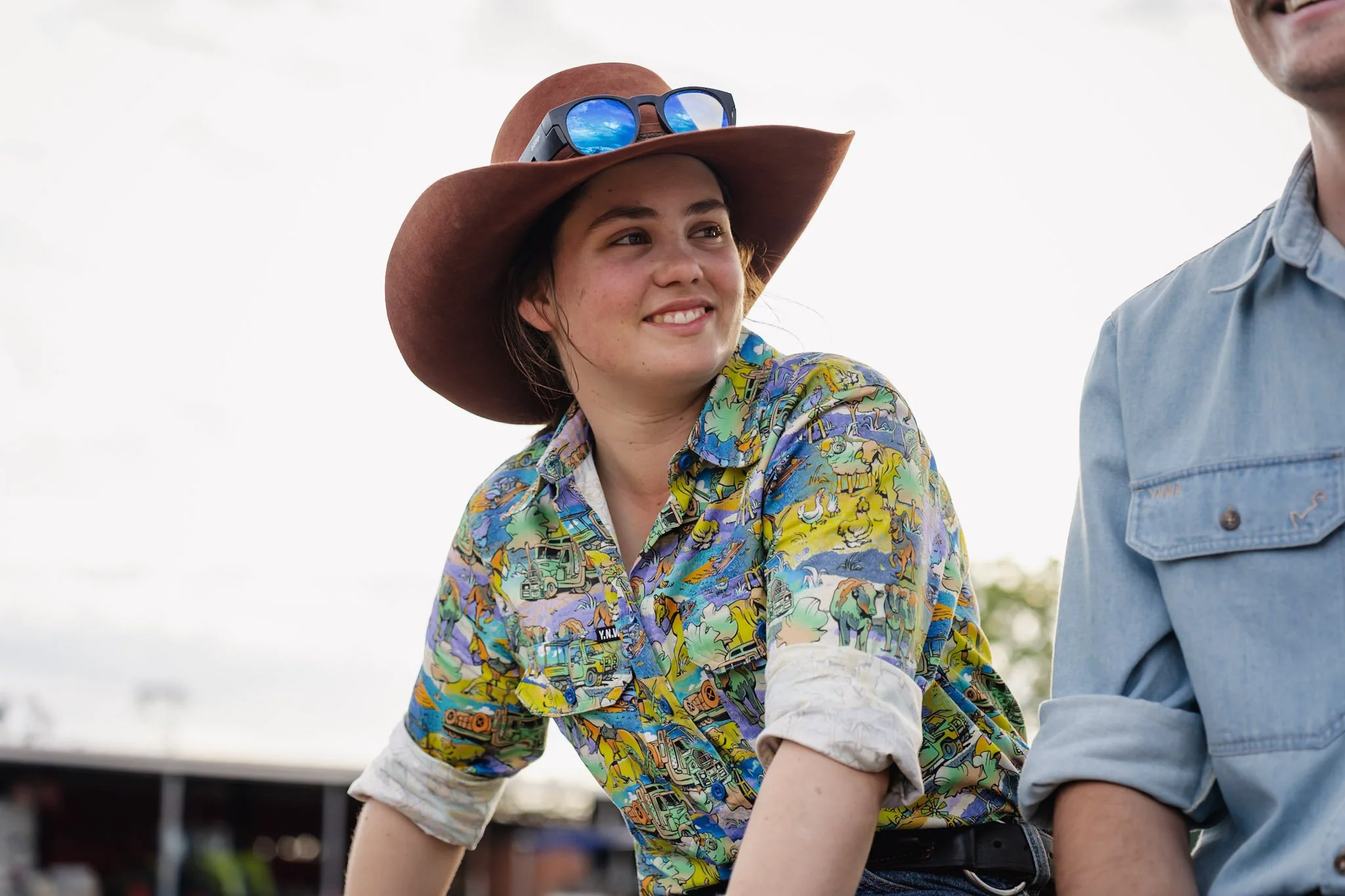Young woman wearing a large brown hat, sunglasses on the hat, and a colorful cowboy shirt, smiling outdoors.