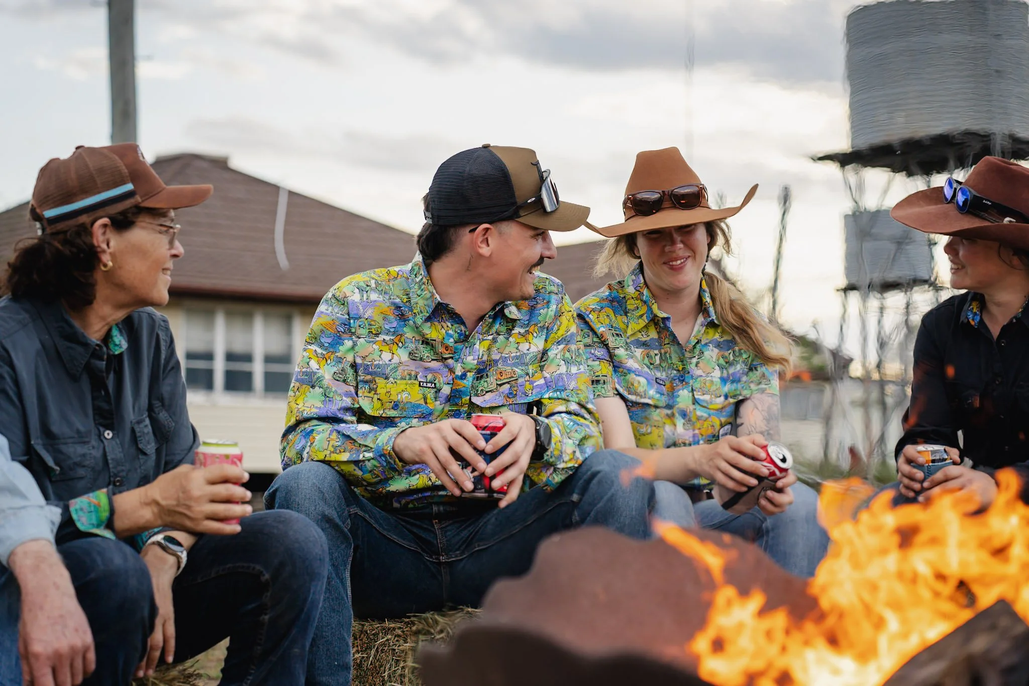Group of five people sitting around a campfire, smiling and talking, with some wearing cowboy hats, in an outdoor setting during sunset or dusk.