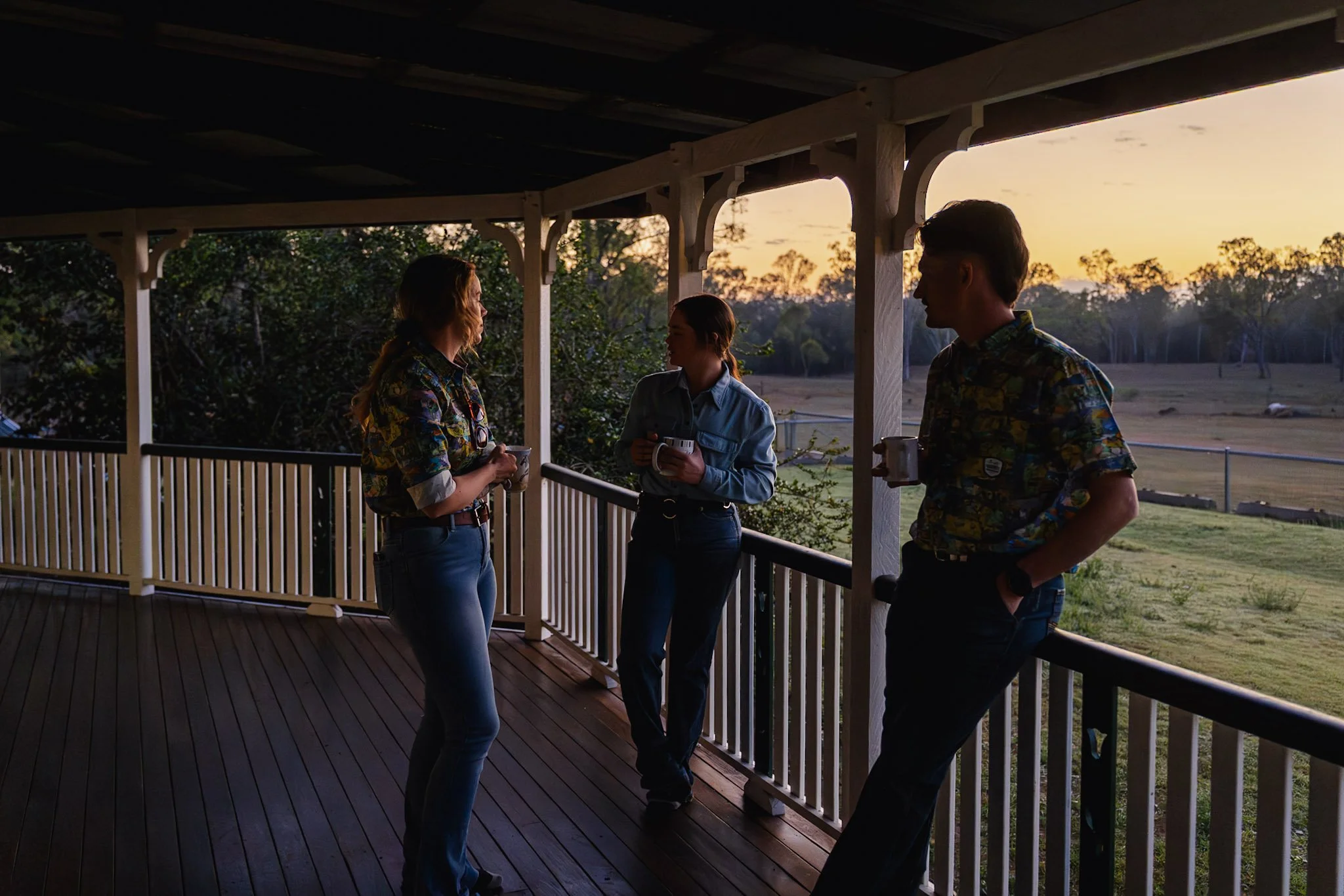 Three people standing on a balcony during sunset, holding mugs and having a conversation. The view shows a grassy field with trees in the background.