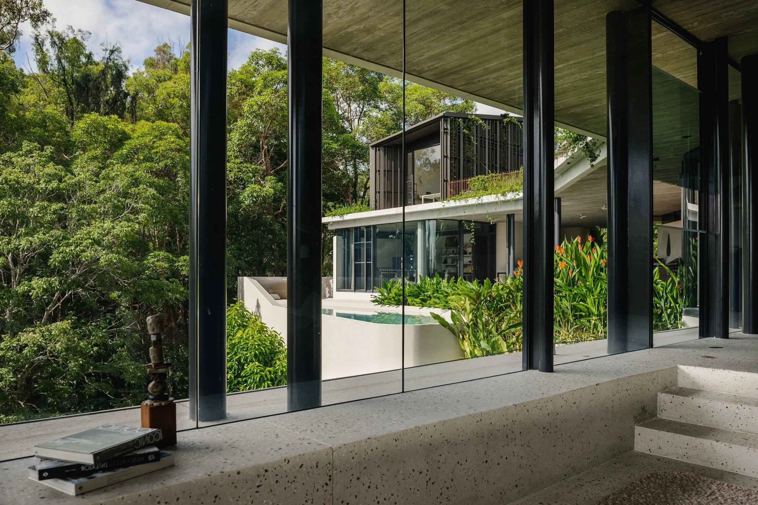 An Architecture photograph showing a modern house with floor-to-ceiling windows, surrounded by lush greenery, seen from an interior view.