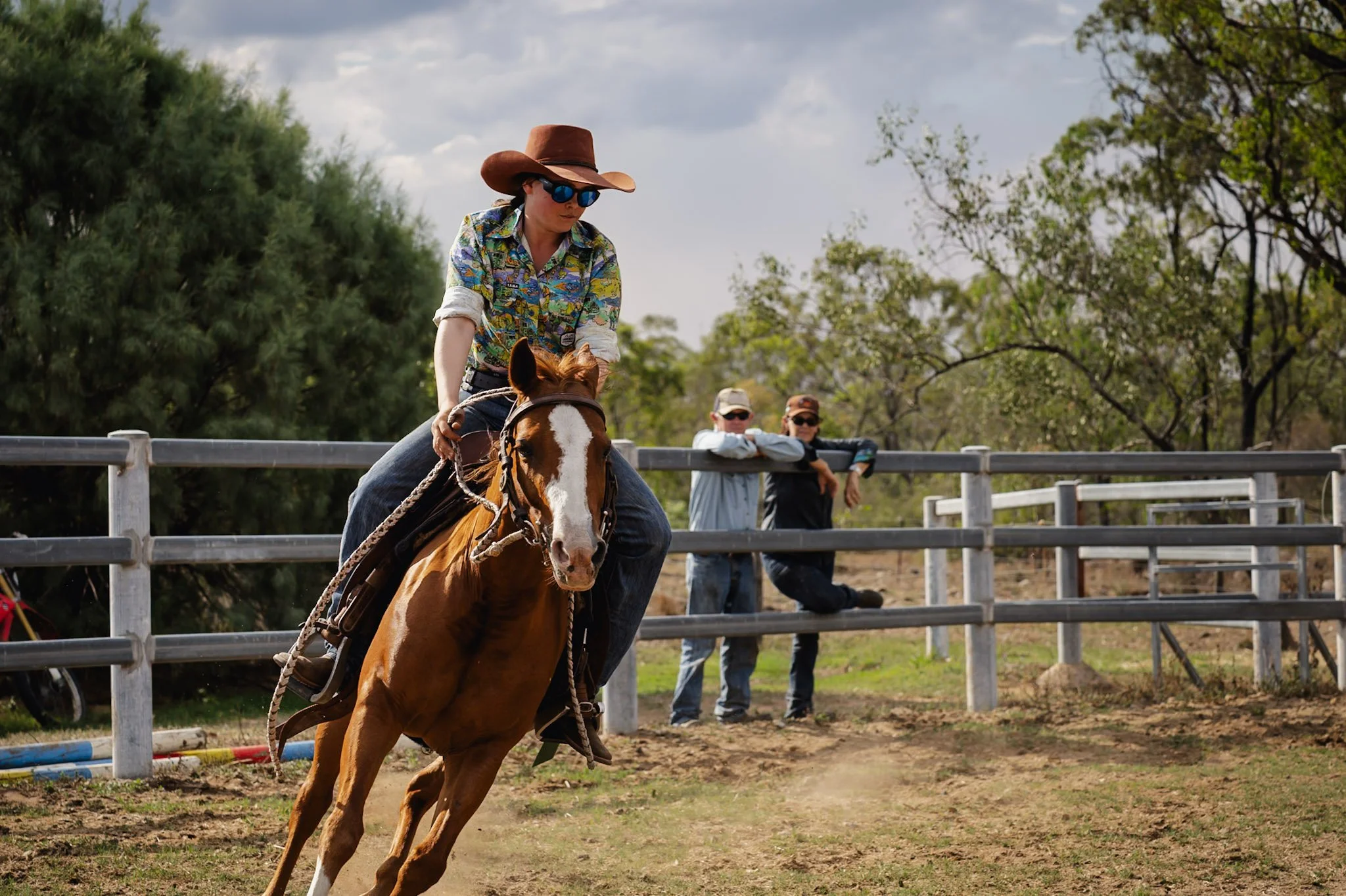 A woman wearing a cowboy hat, sunglasses, and a colorful shirt rides a brown and white horse on a dirt track with two men watching from behind a white fence.