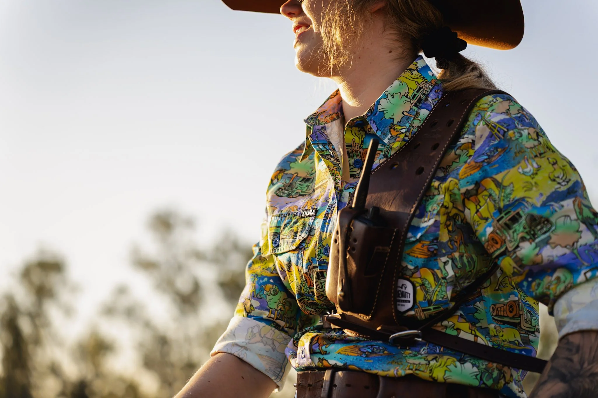 Close-up of a woman in colorful western-style shirt and cowboy hat, with a brown leather holster on her chest, outdoors at sunset.