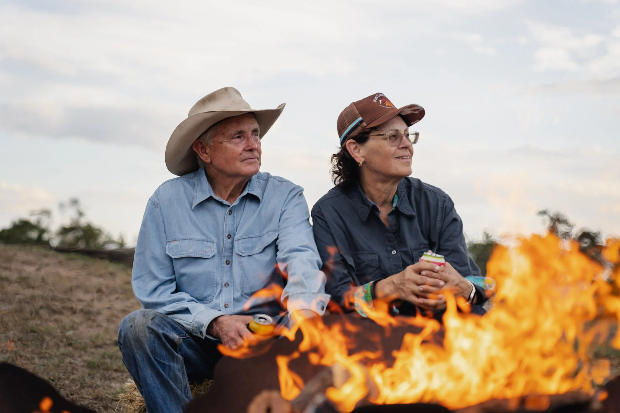 An elderly man and woman sitting outdoors near a campfire during the evening. The man wears a cowboy hat and a light denim shirt, while the woman wears glasses, a brown cap, and a dark shirt. The woman holds a drink and they both look thoughtful.