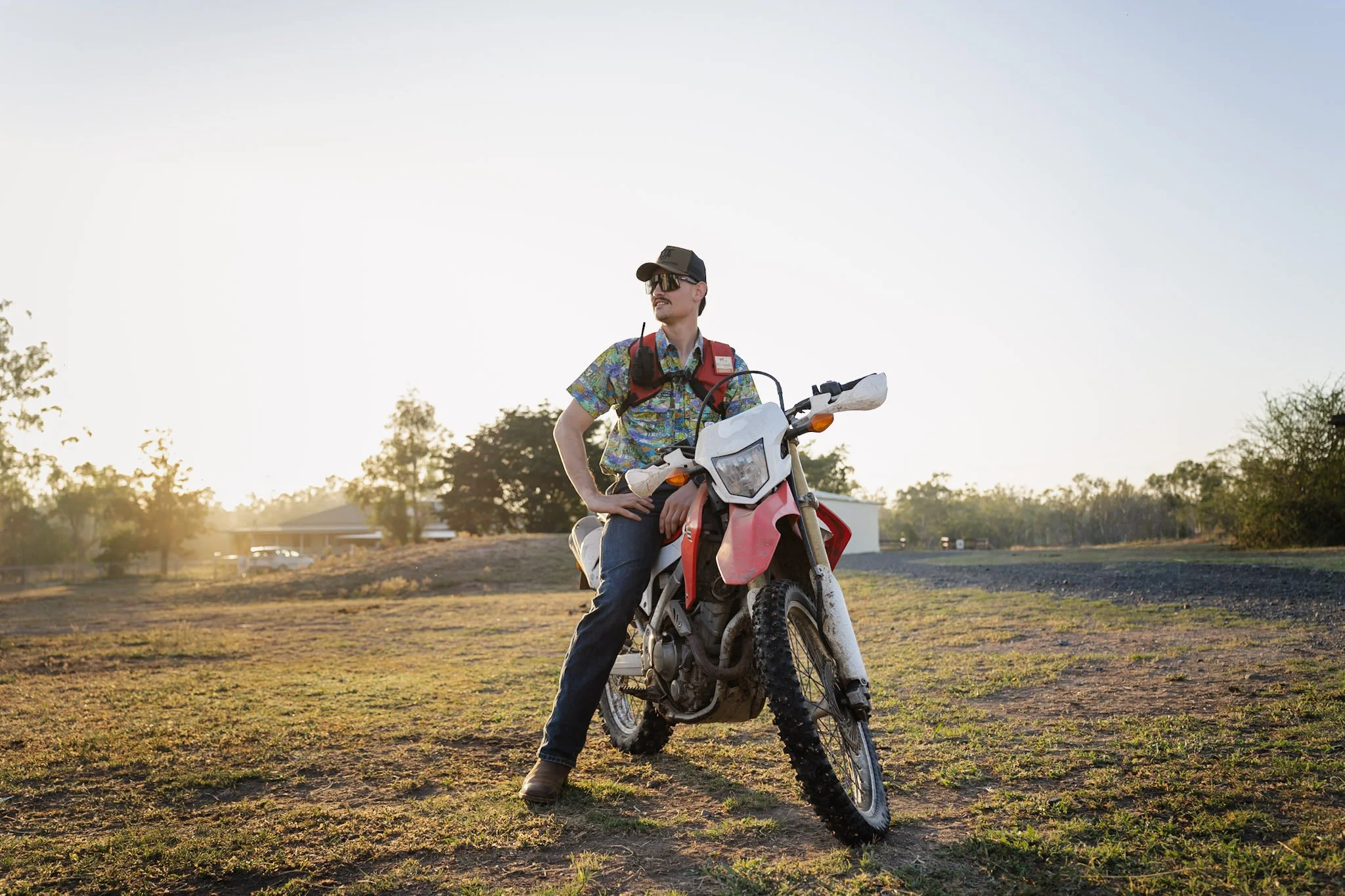 A man standing next to a dirt bike in a grassy outdoor area at sunset, wearing casual clothes, a cap, sunglasses, and a backpack.