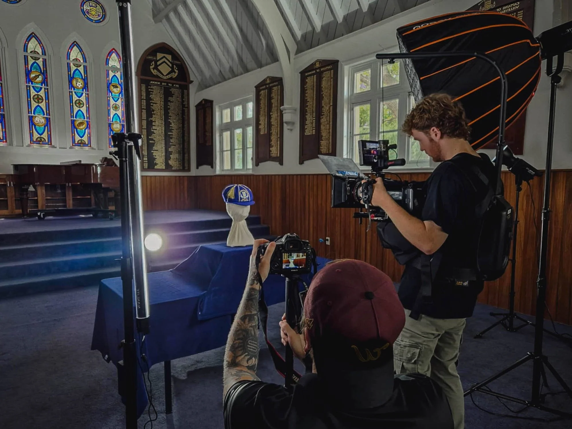 Camera Crew setting up a video camera and lighting equipment in a chapel with stained glass windows and wooden paneling, focusing on a cap placed on a table.