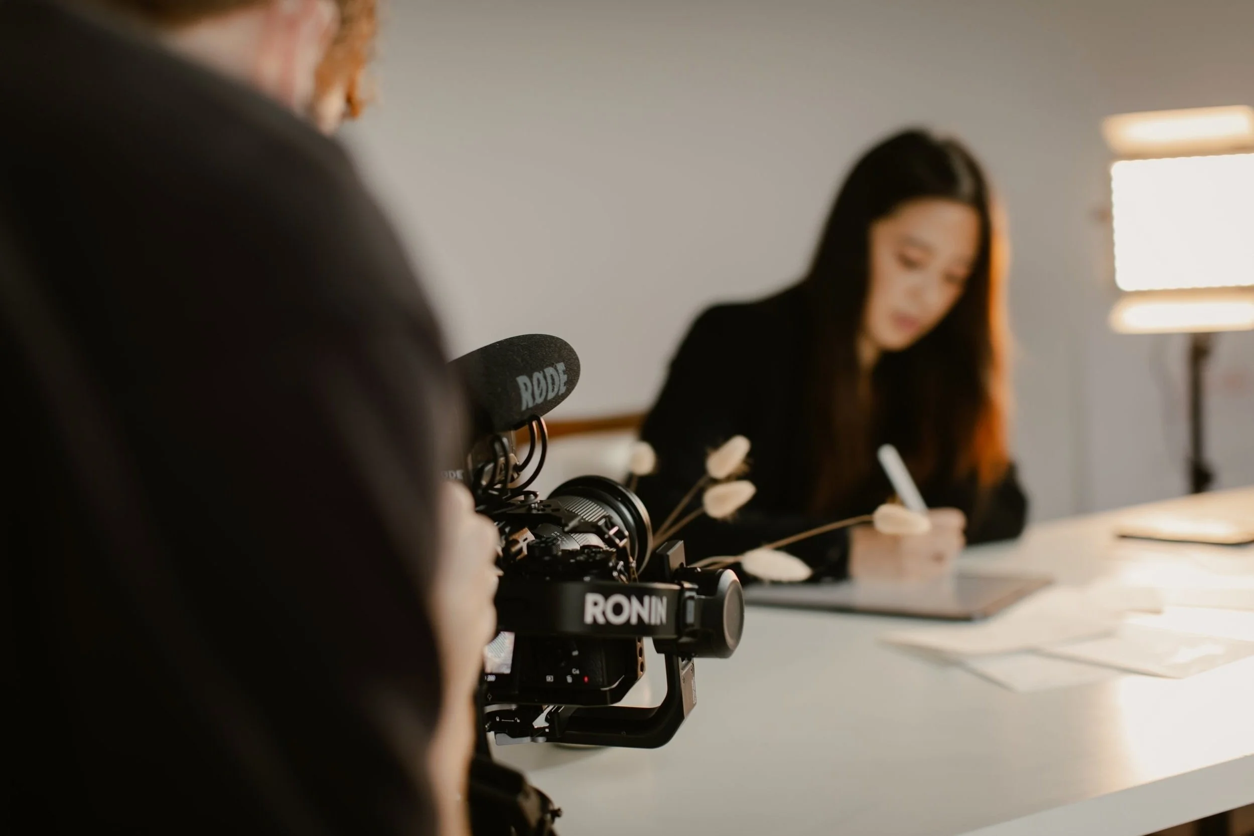 A videographer films a person writing at a desk for social media content, with a video camera setup featuring a RODE microphone and RONIN stabilizer.