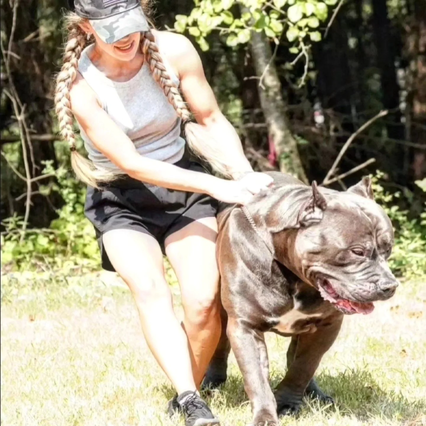 Woman with long braided hair riding on the back of a large, muscular dog in a grassy outdoor area.