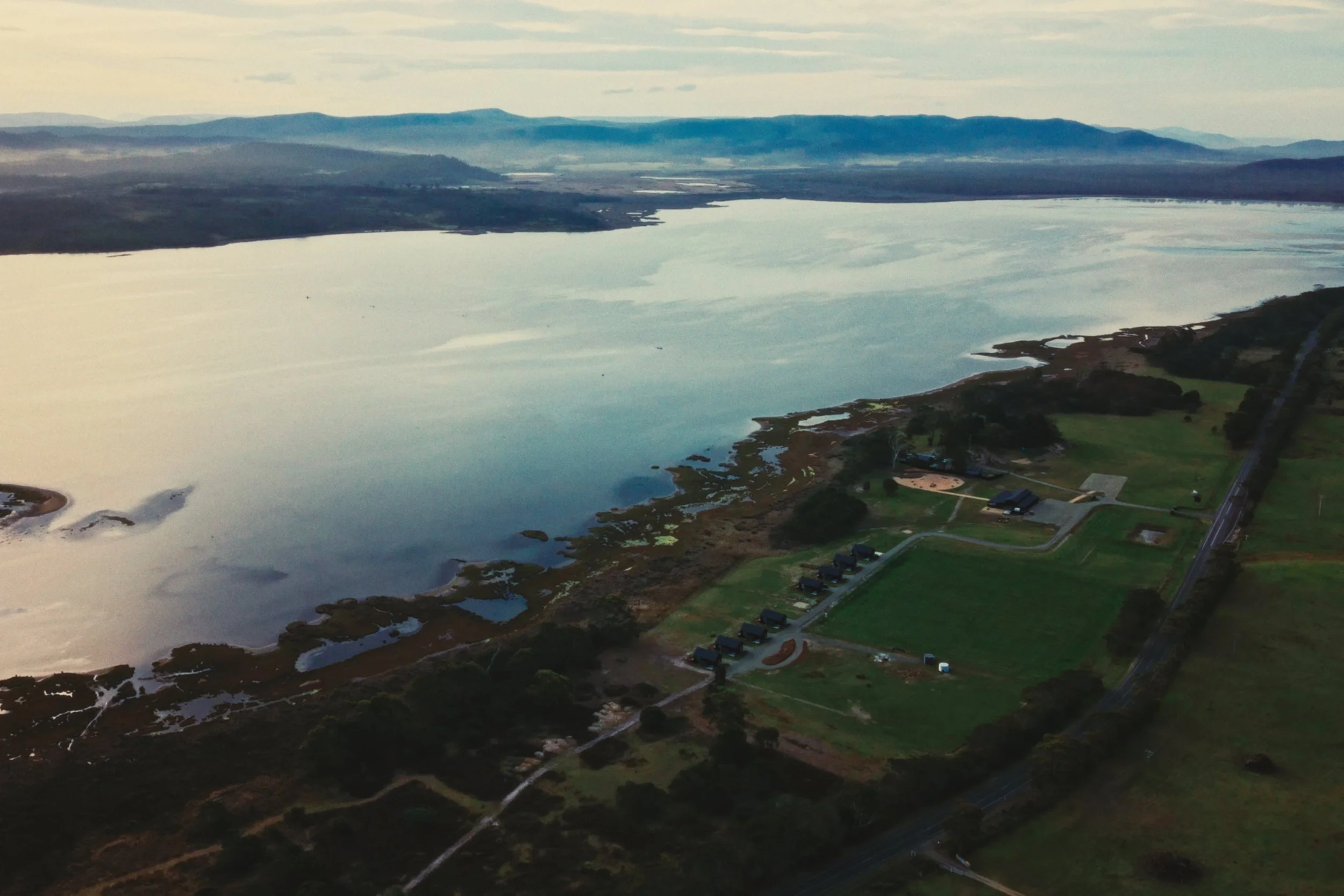 Aerial view of a large body of water with a park and a parking lot along the shoreline, surrounded by green grassy areas and trees, with mountains in the distance under a partly cloudy sky.