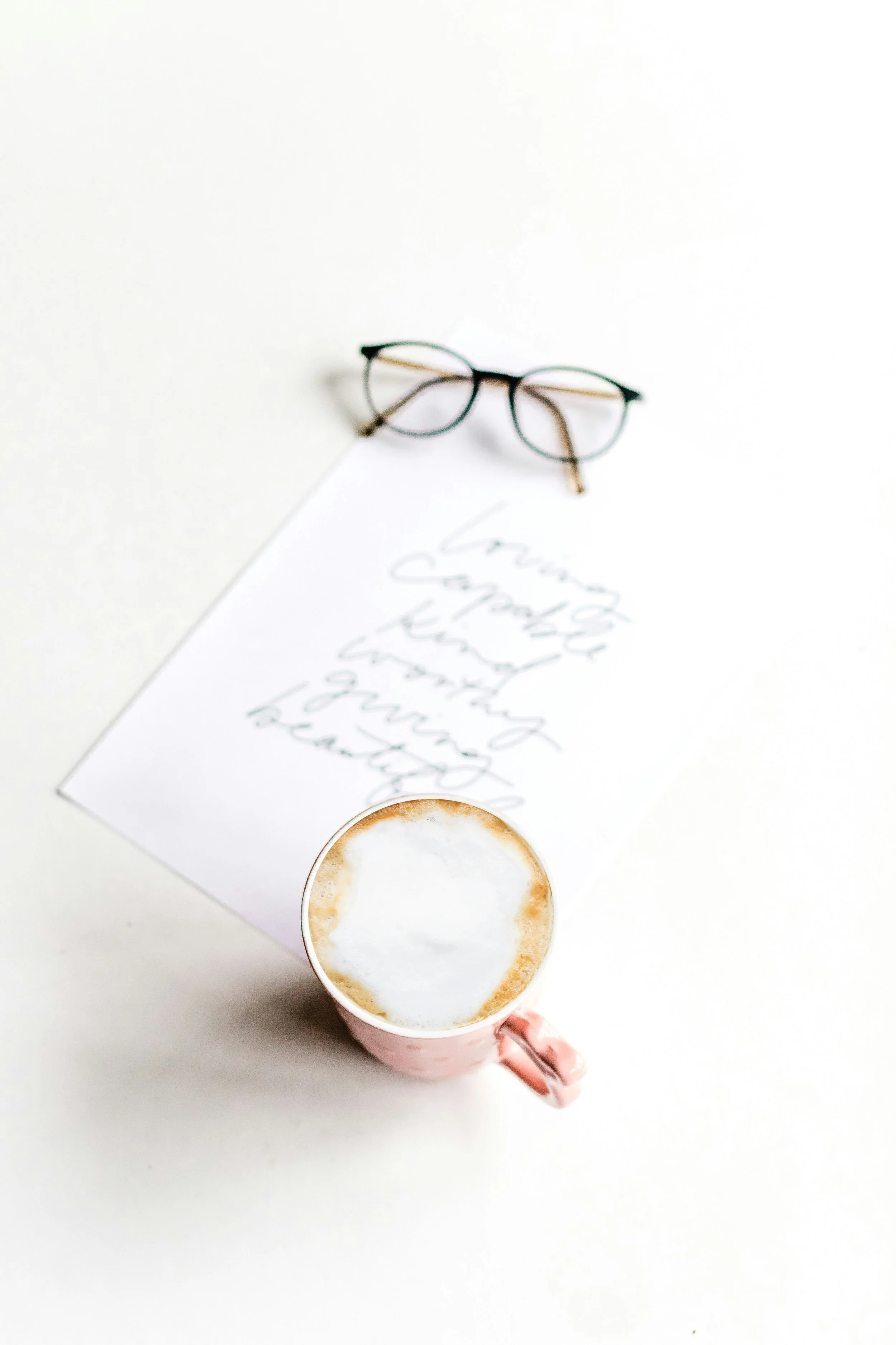 A person holding a pink cup of cappuccino with foam on top, positioned over a white surface. There is a handwritten note and a pair of glasses on a piece of paper nearby.