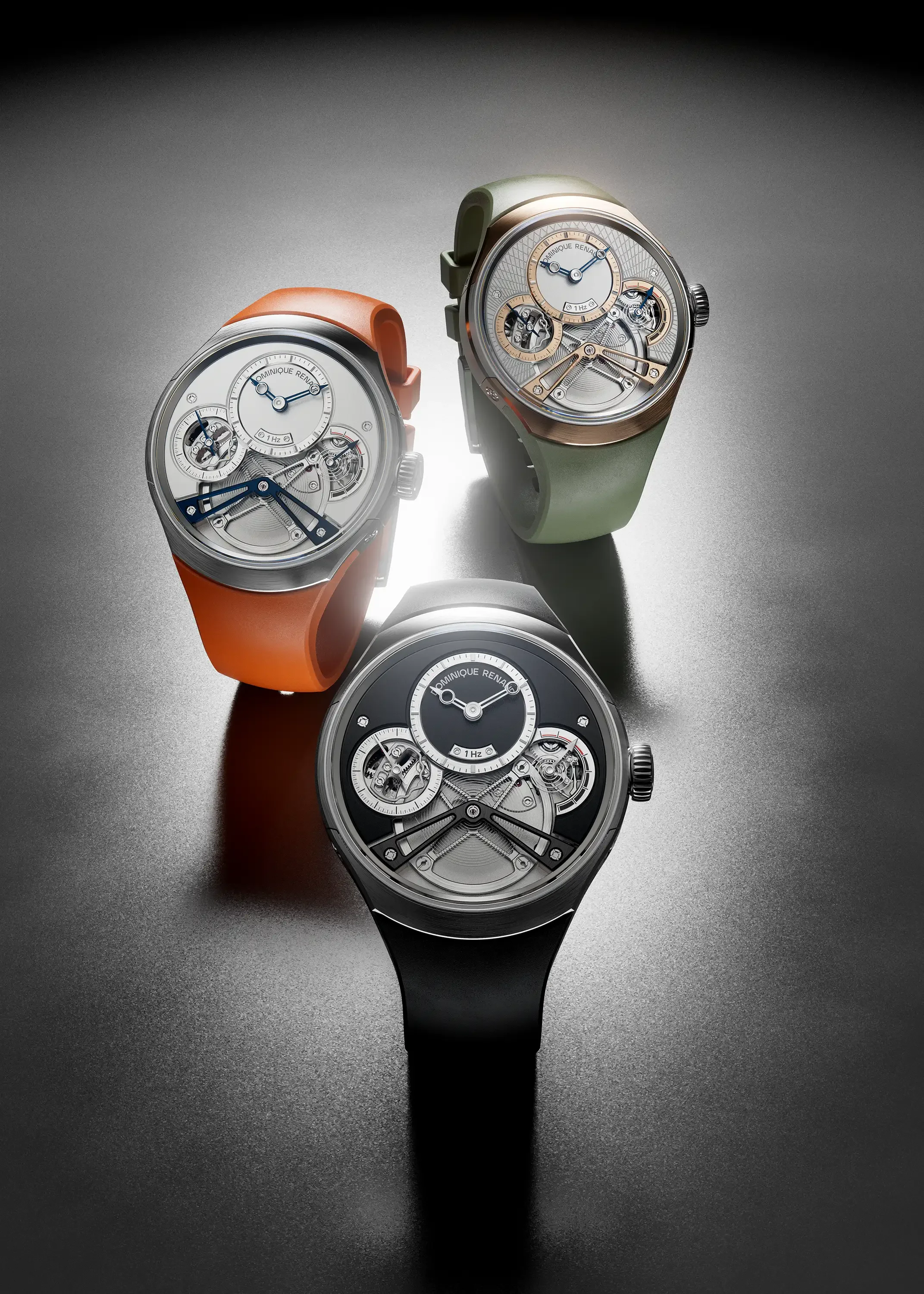 Trois montres avec des cadrans ouverts montrant des mécanismes internes, en différentes couleurs de bracelets, sur une surface sombre avec un éclairage centré.