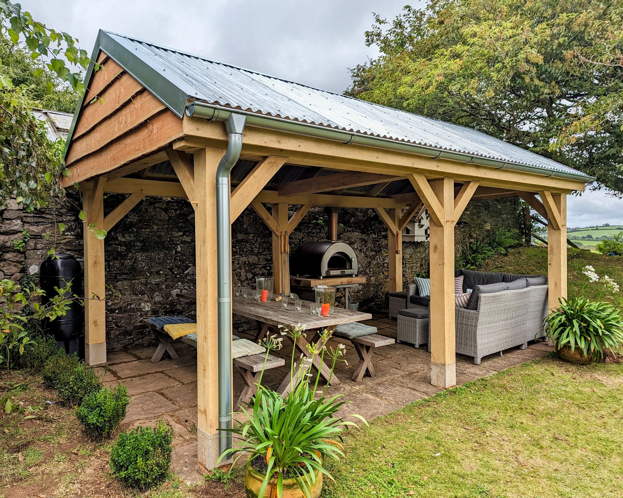 AGreen oak pergola structure over an outdoor living area with seating, dining benches and a pizza oven in a Devon garden.