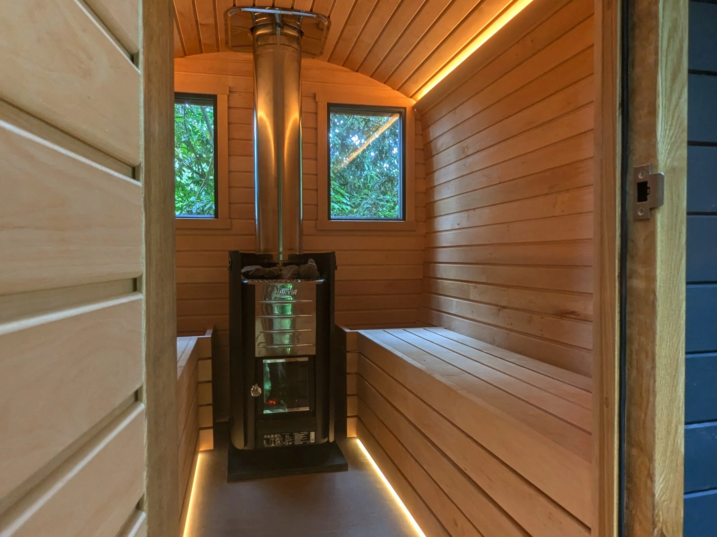 A small wooden sauna room with a wood stove and windows showing green foliage outside.