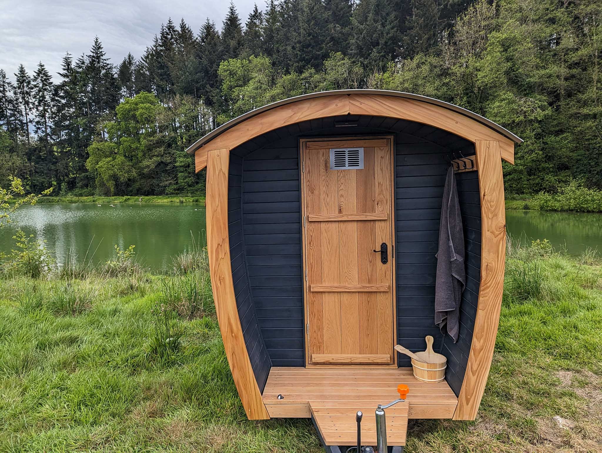 A small, portable outdoor sauna with a wooden door and black siding, situated by a lake with trees in the background.