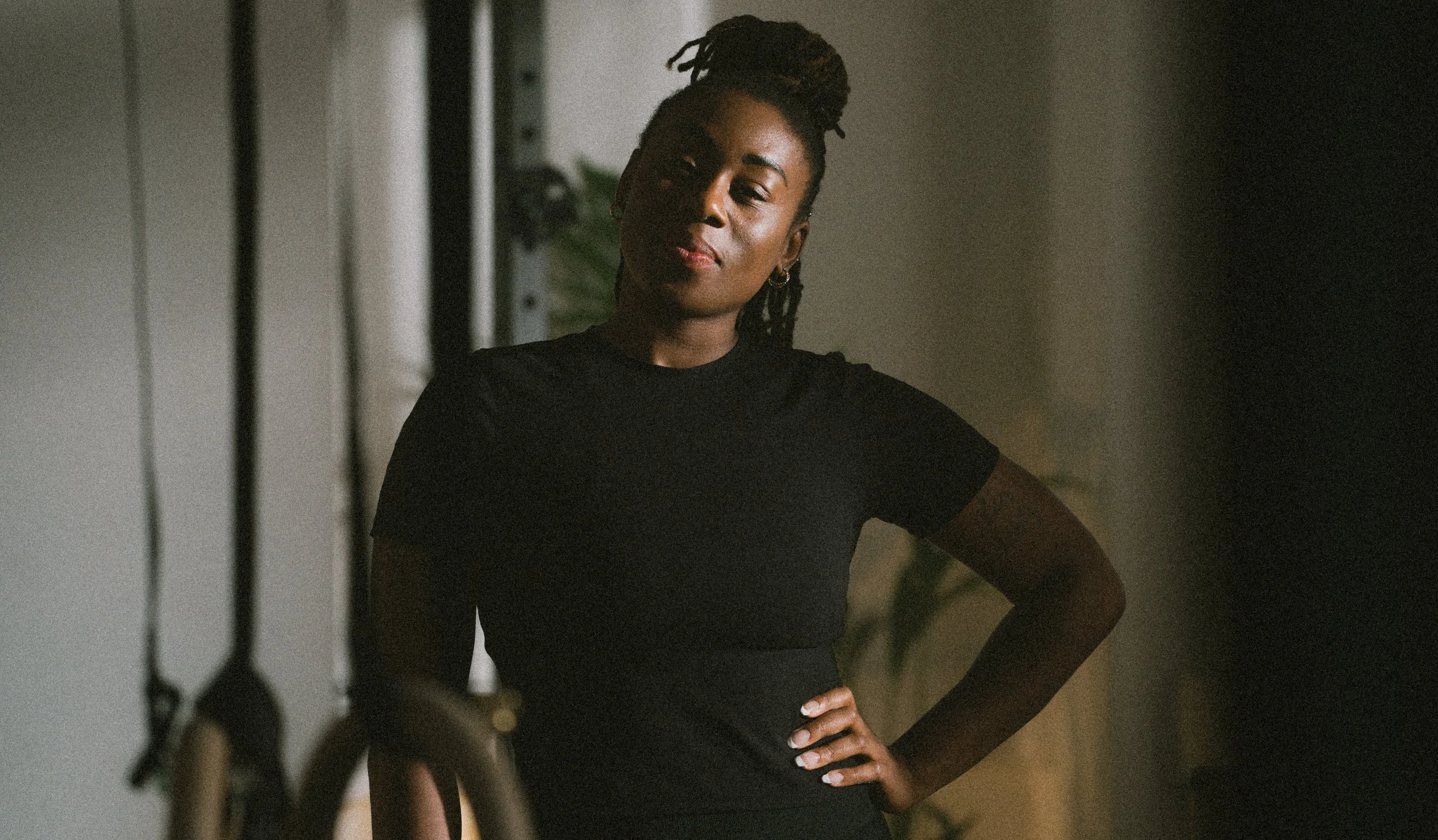 A woman with braided hair styled in an updo wearing a black t-shirt standing indoors with her hand on her hip, looking at the camera with a neutral expression.