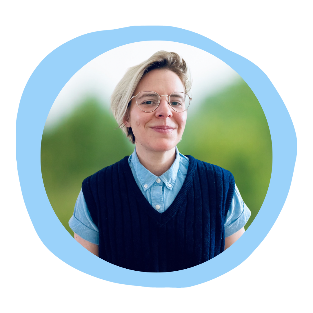 Portrait of a young woman with short blonde hair, glasses, wearing a light blue shirt and dark blue sleeveless sweater, standing outdoors with greenery in the background.