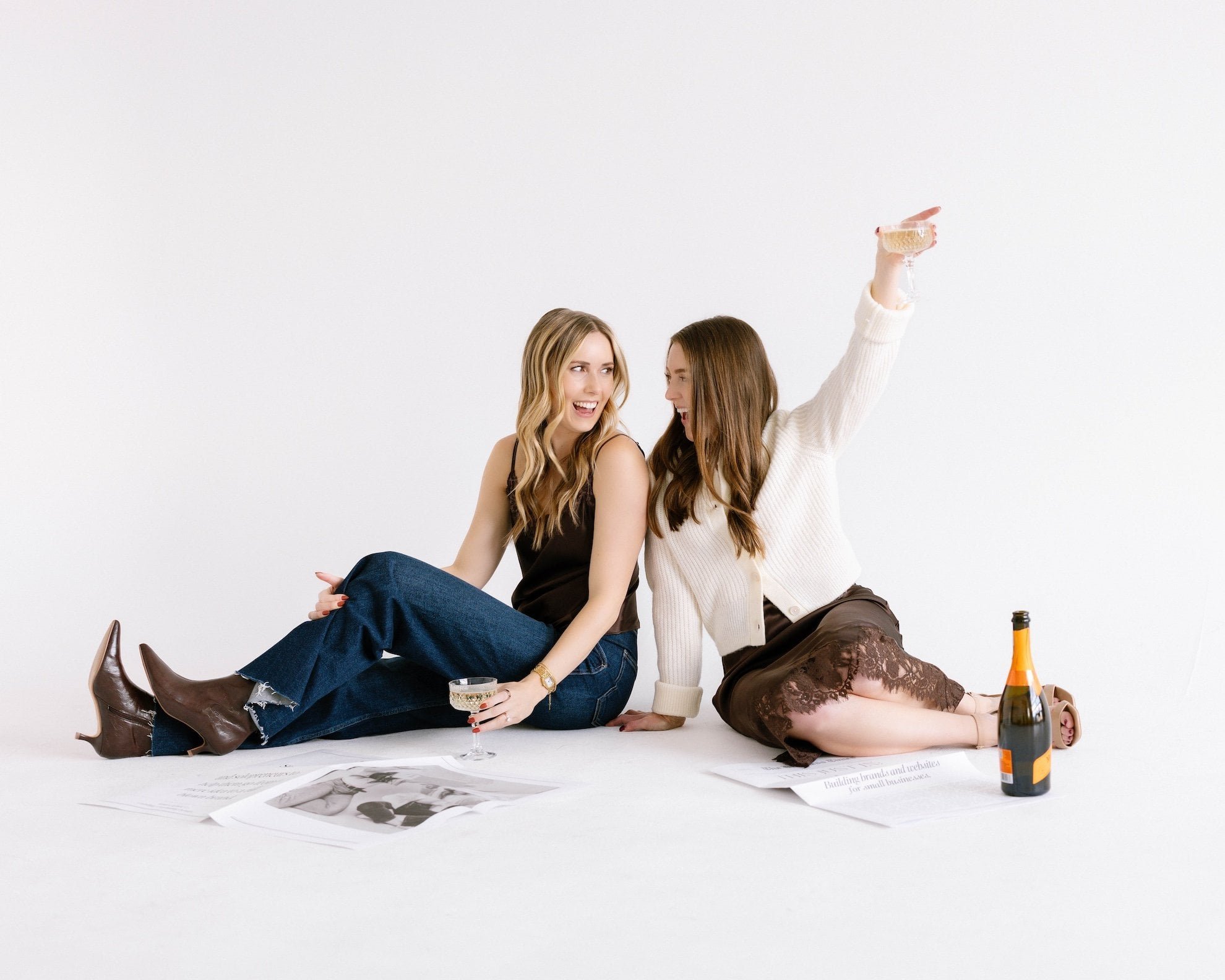 Two women sitting on the floor against a plain white background, smiling and engaging with each other. One woman has blonde hair and is wearing a black top with blue jeans and boots, holding a wine glass. The other woman has brown hair and is wearing a cream sweater with a lace skirt, positioned on her knees. There are scattered papers, a bottle of wine, and a wine glass on the floor nearby.