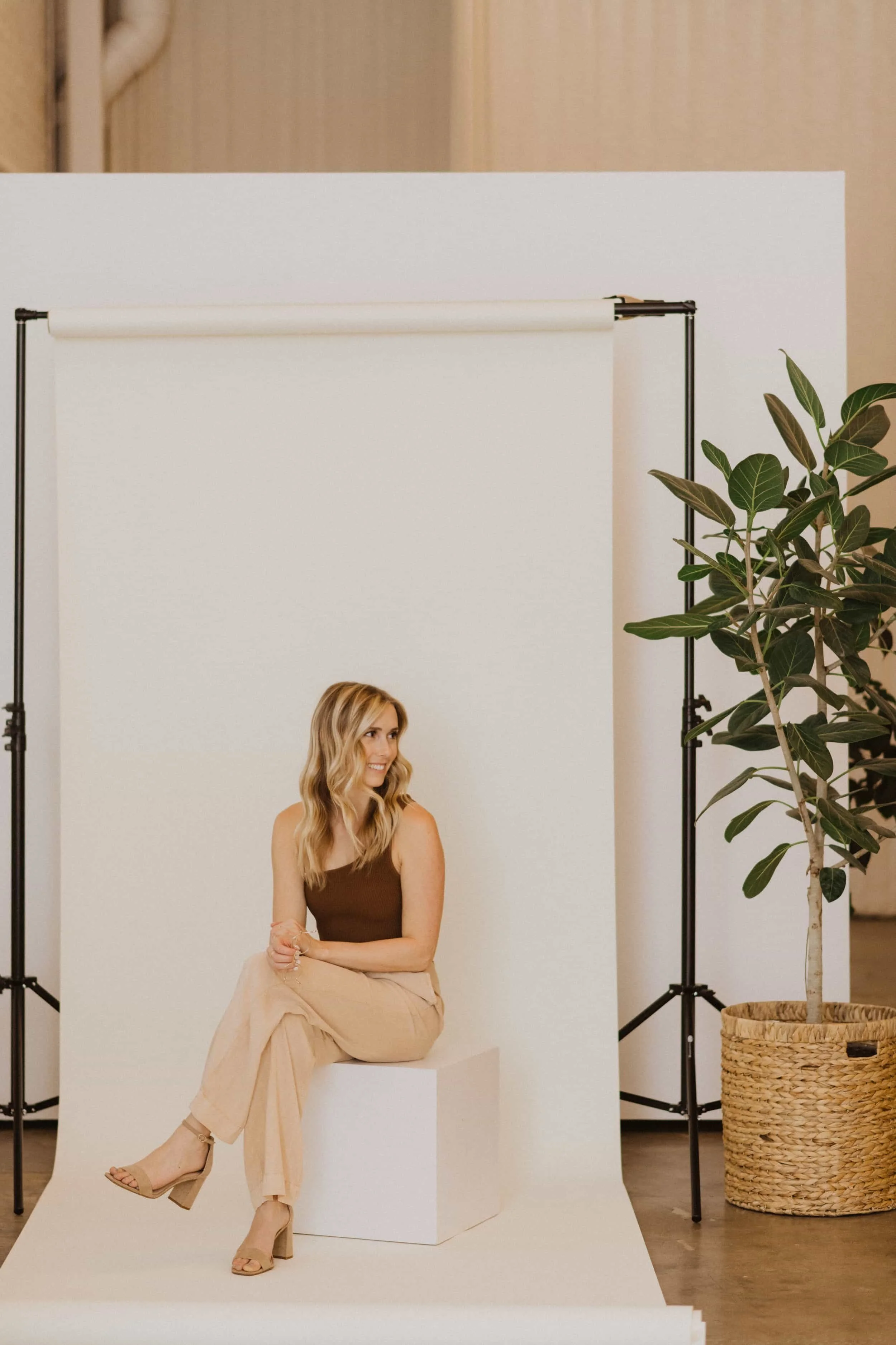 A woman with long blonde hair in a brown tank top and beige wide-leg pants sitting on a white platform, smiling and looking to her right, in front of a white photography backdrop with a green potted plant nearby.
