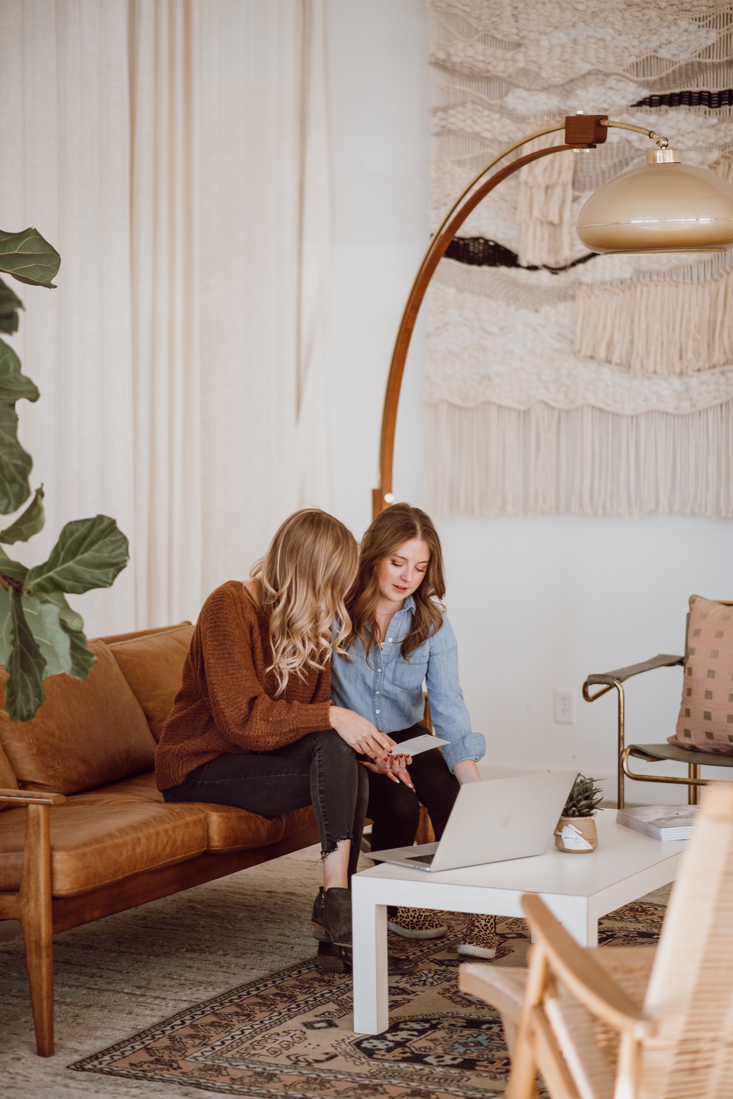 Two women sitting on a leather couch looking at a laptop in a cozy living room with a woven wall hanging and mid-century decor.
