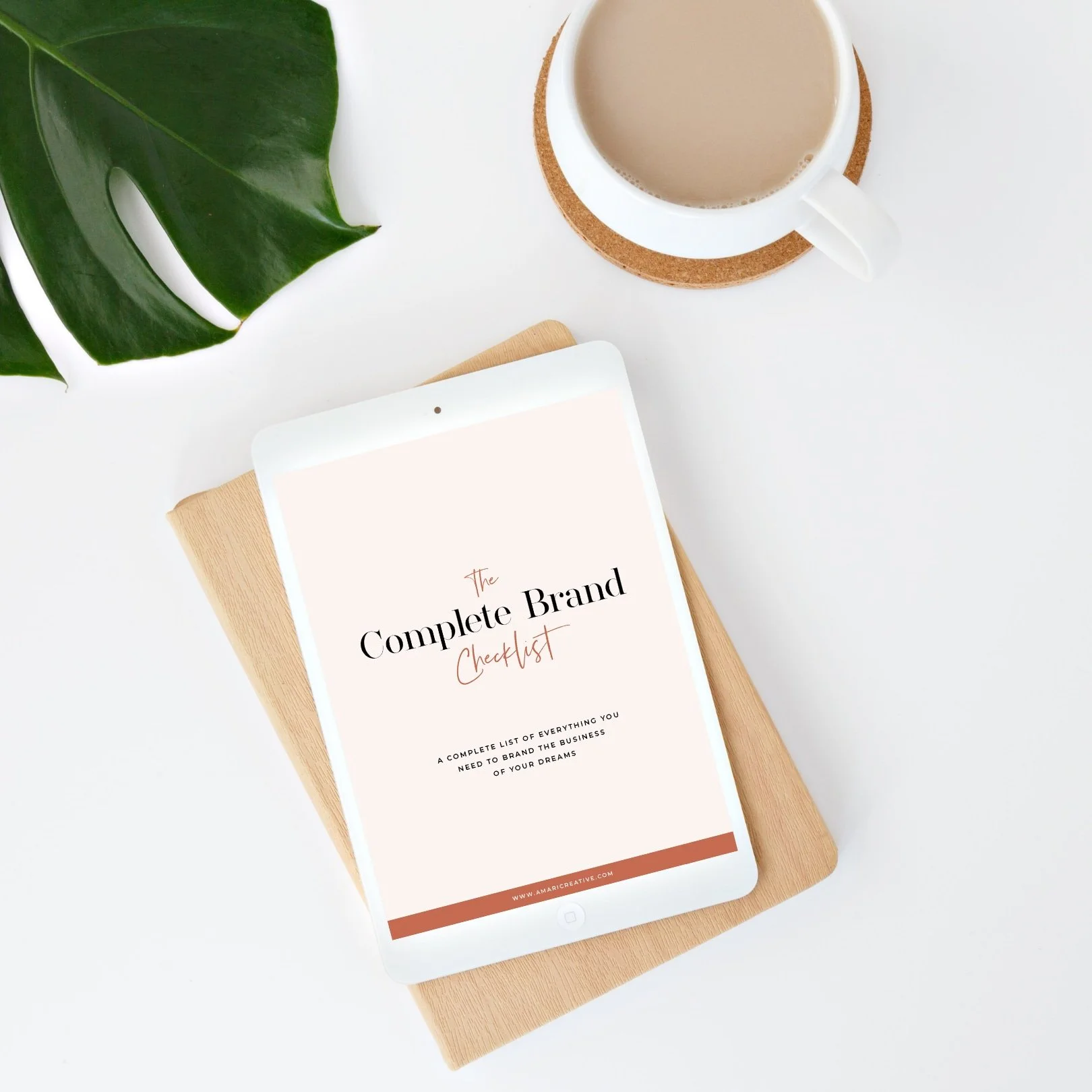 A white tablet displaying the cover of 'The Complete Brand Checklist' on top of a closed light brown notebook, a white coffee cup with a cork coaster, and a large green leaf on a white surface.