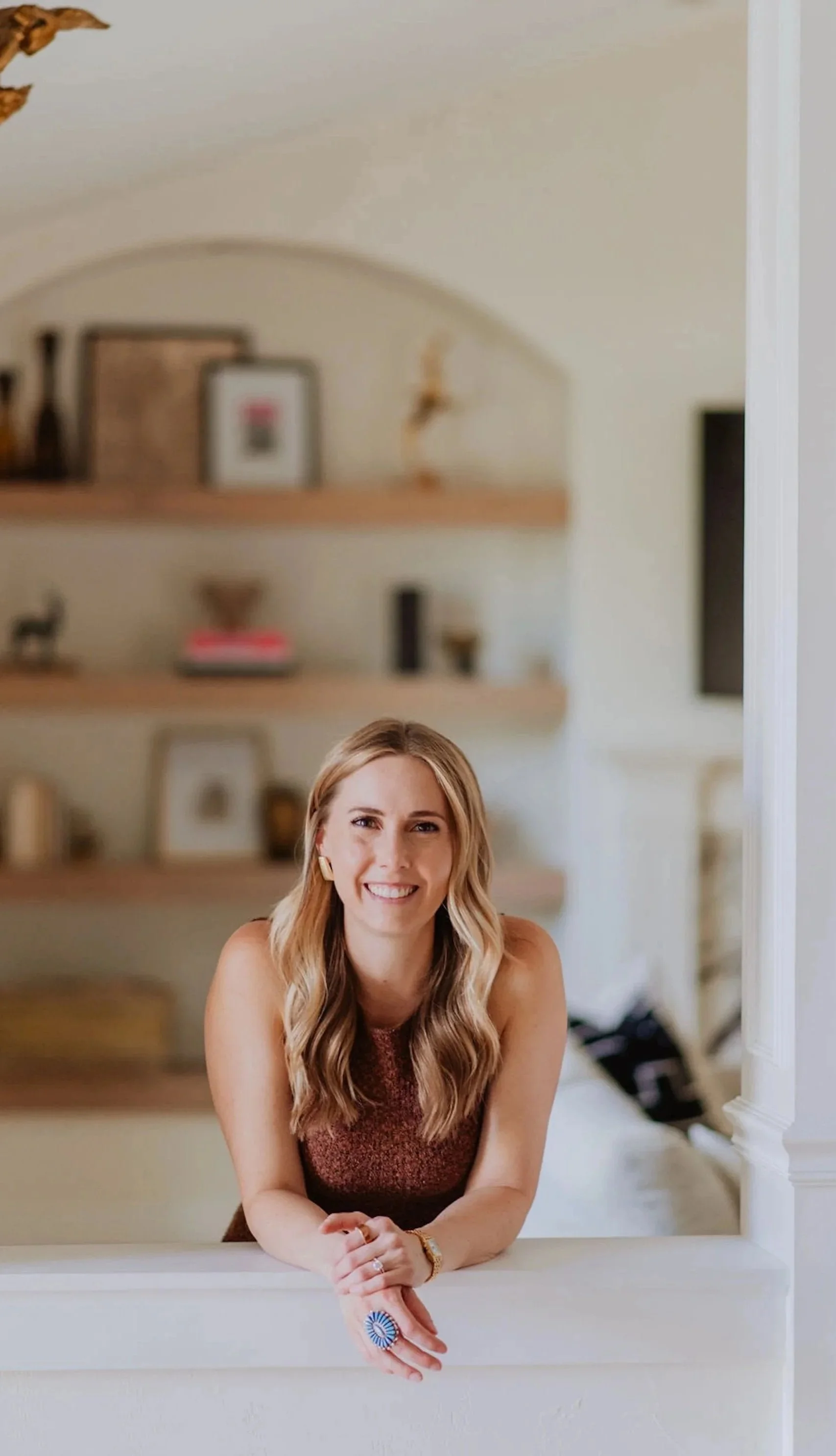 A woman smiling and sitting at a white table in a cozy room with wooden shelves and decorative items in the background.