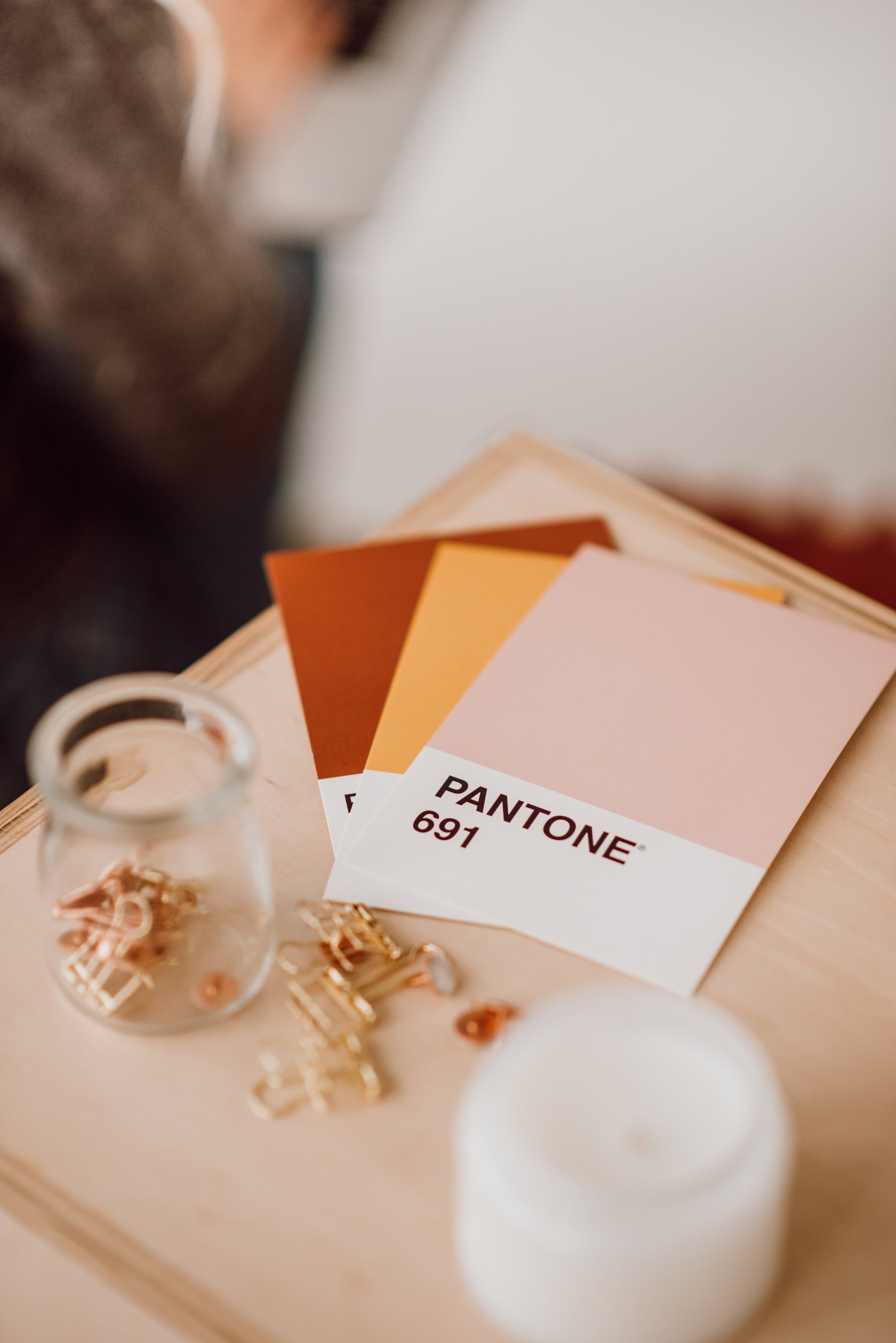 Pantone color swatches with the label 'Pantone 691' on a white card, a small glass jar, and scattered gold paper clips on a light-colored surface.