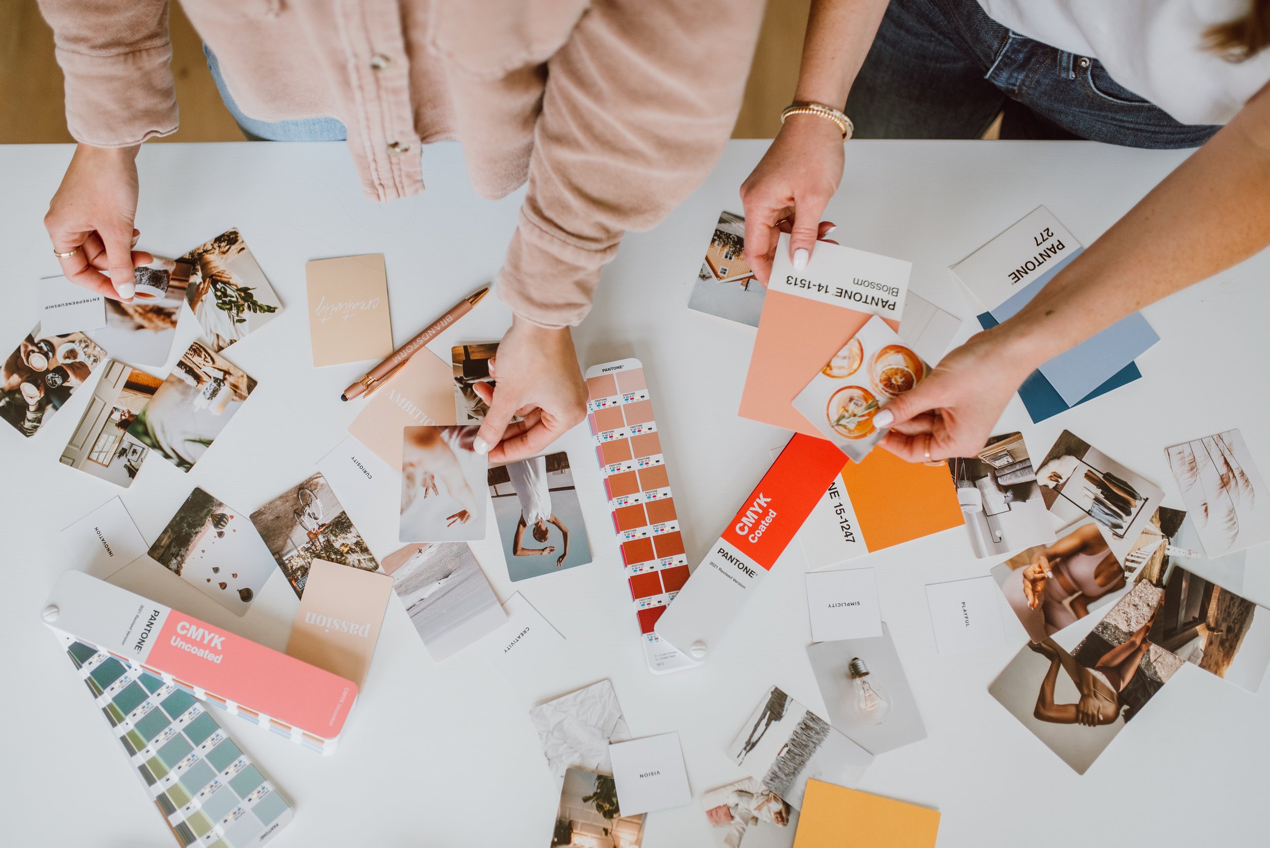 Overhead view of two people at a white table discussing and selecting paint colors using color swatches, sample photos, and paint samples with a focus on the color palette and design ideas.
