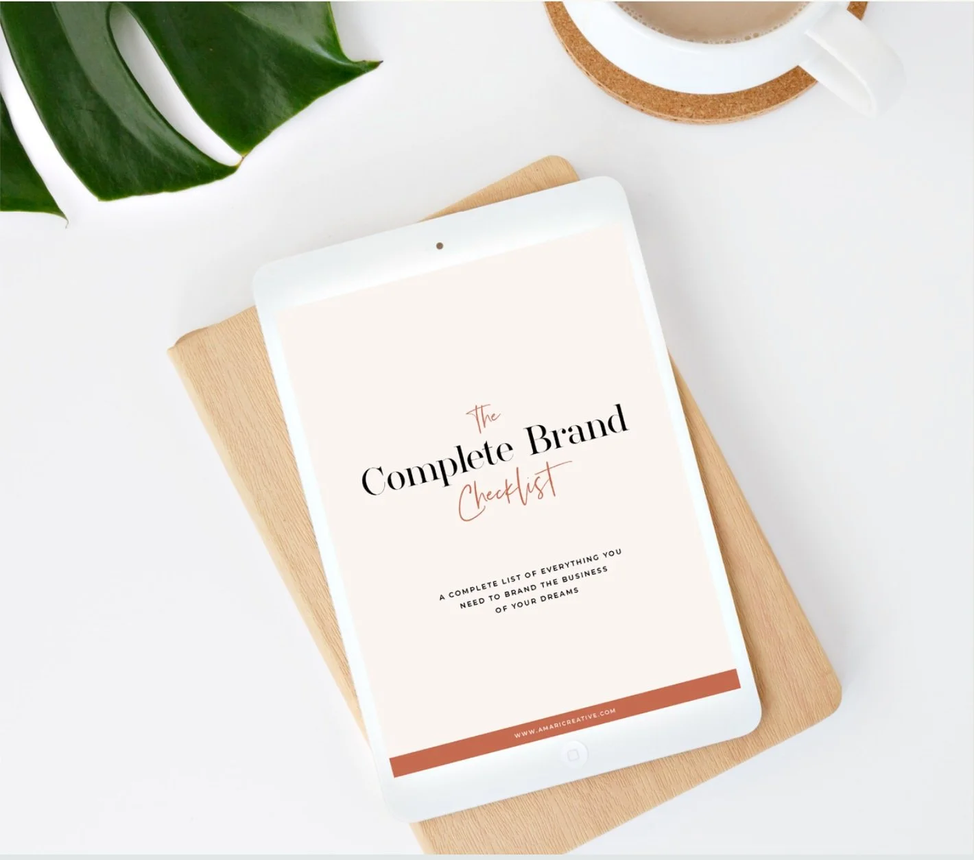 A digital tablet displaying a booklet titled 'The Complete Brand Checklist' resting on a wooden clipboard, with a green leaf and a cup of coffee nearby on a white surface.
