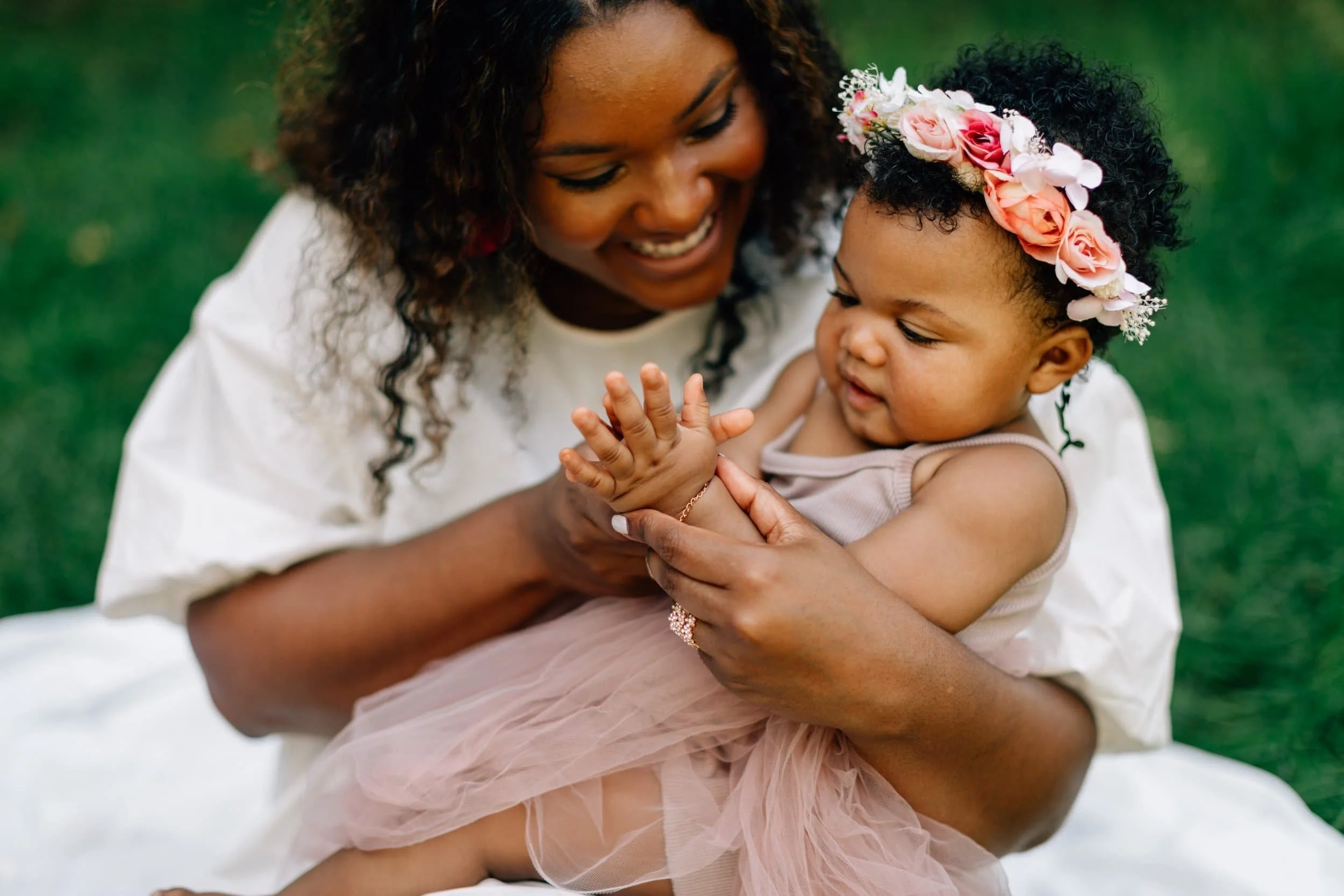 Smiling woman in white holds a baby in a pink dress and flower crown while sitting on grass.