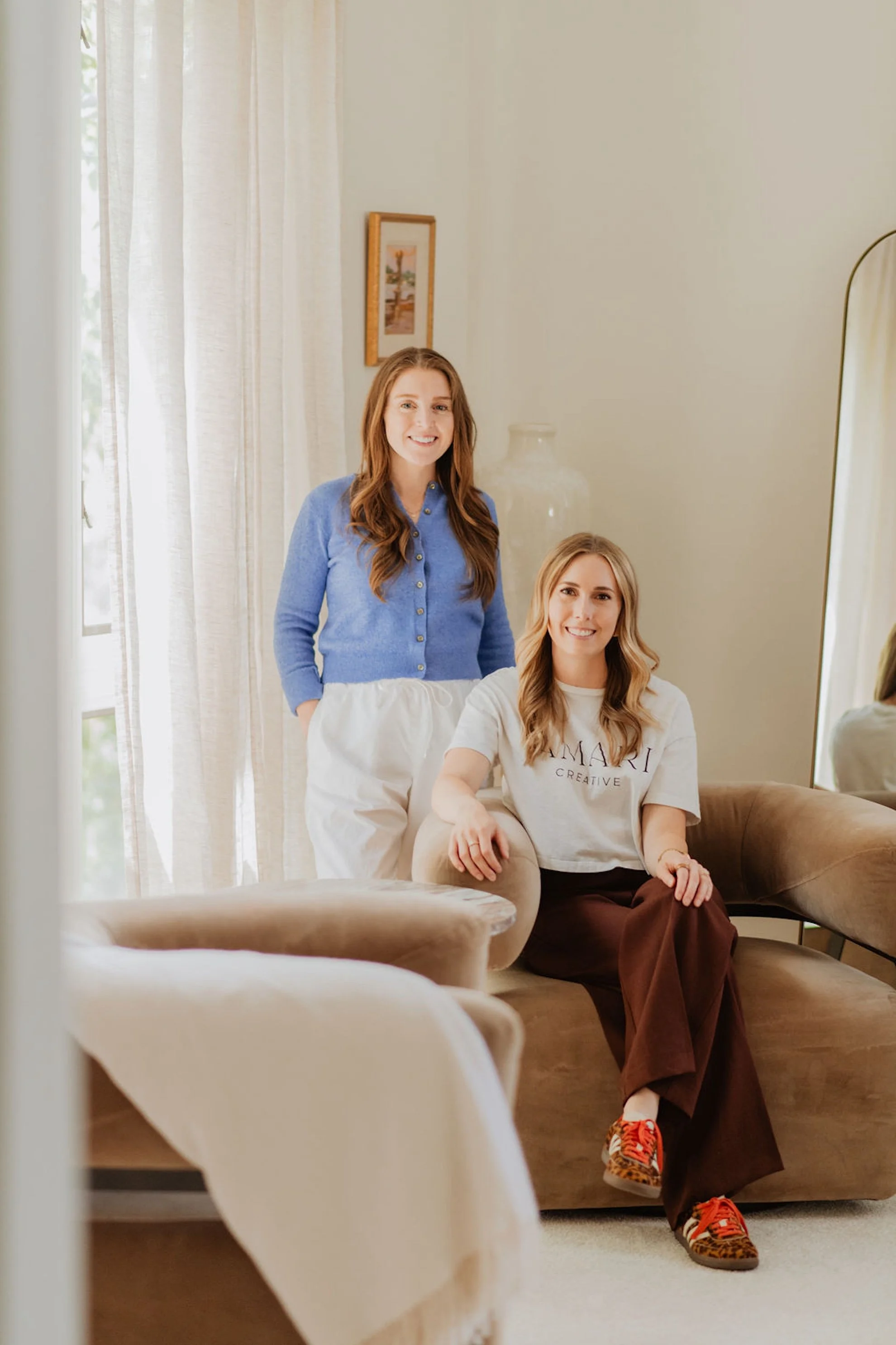 Two women sitting and standing in a bright room near a large mirror, with neutral-colored walls, curtains, and a framed artwork on the wall.