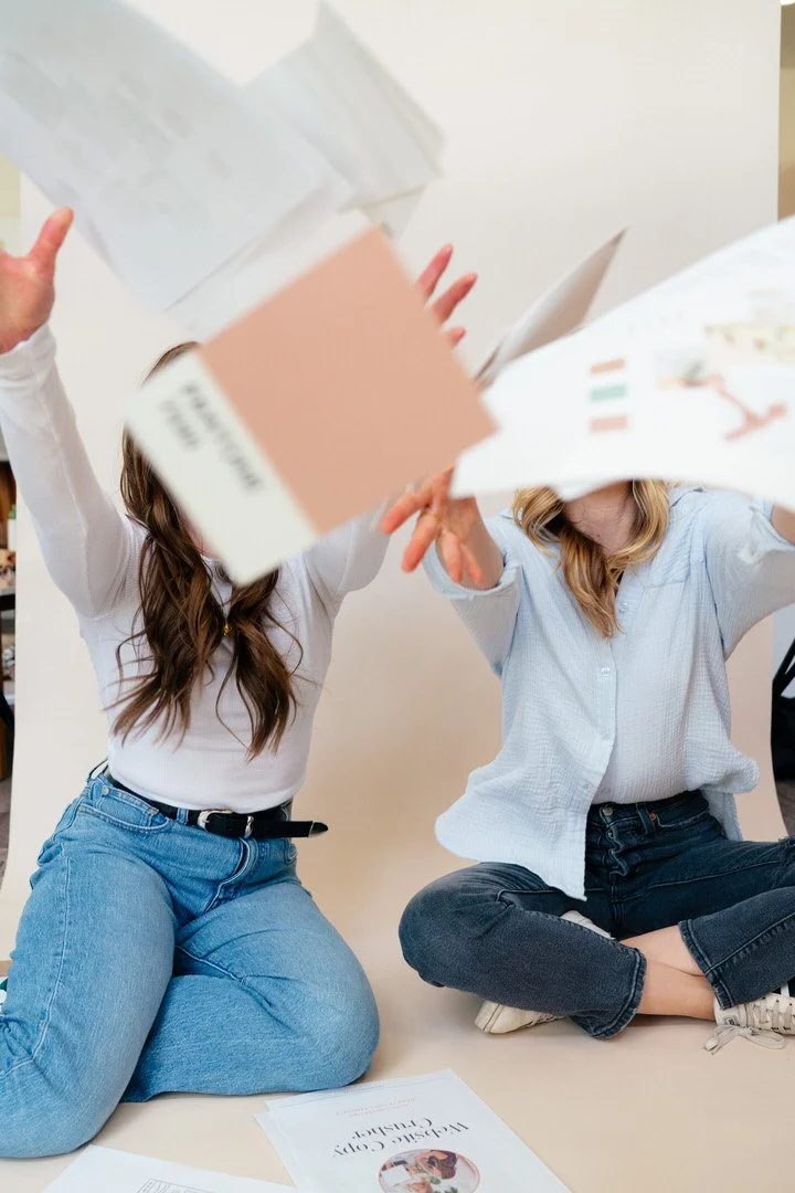 Two women sitting on the floor, tossing papers and books into the air, in a playful manner.