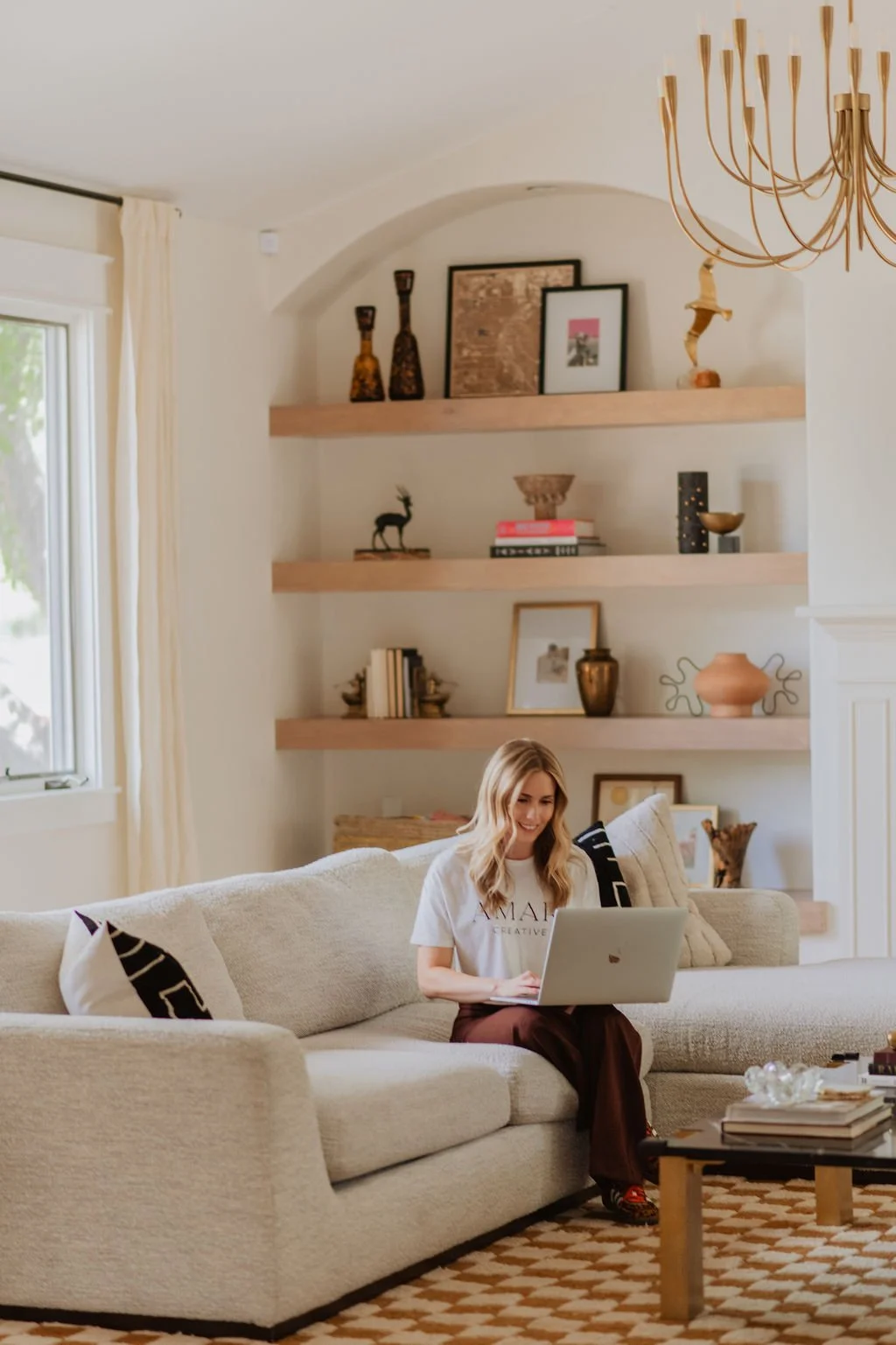 A woman sitting on a beige sofa using a laptop in a cozy living room with shelves decorated with art, books, and decorative objects.