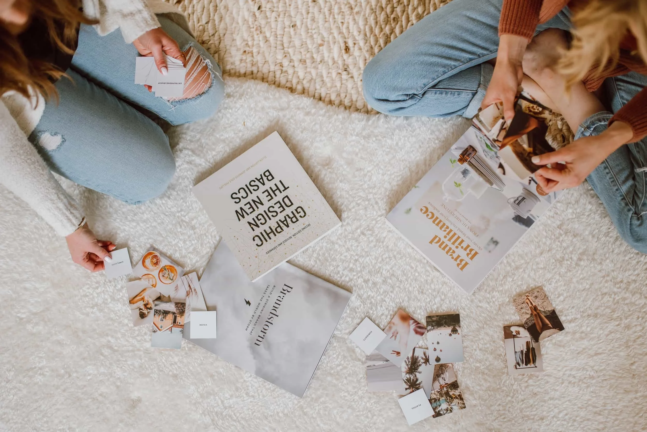 Two women sitting on a beige rug, sorting through magazines, brochures, and business cards on the floor.