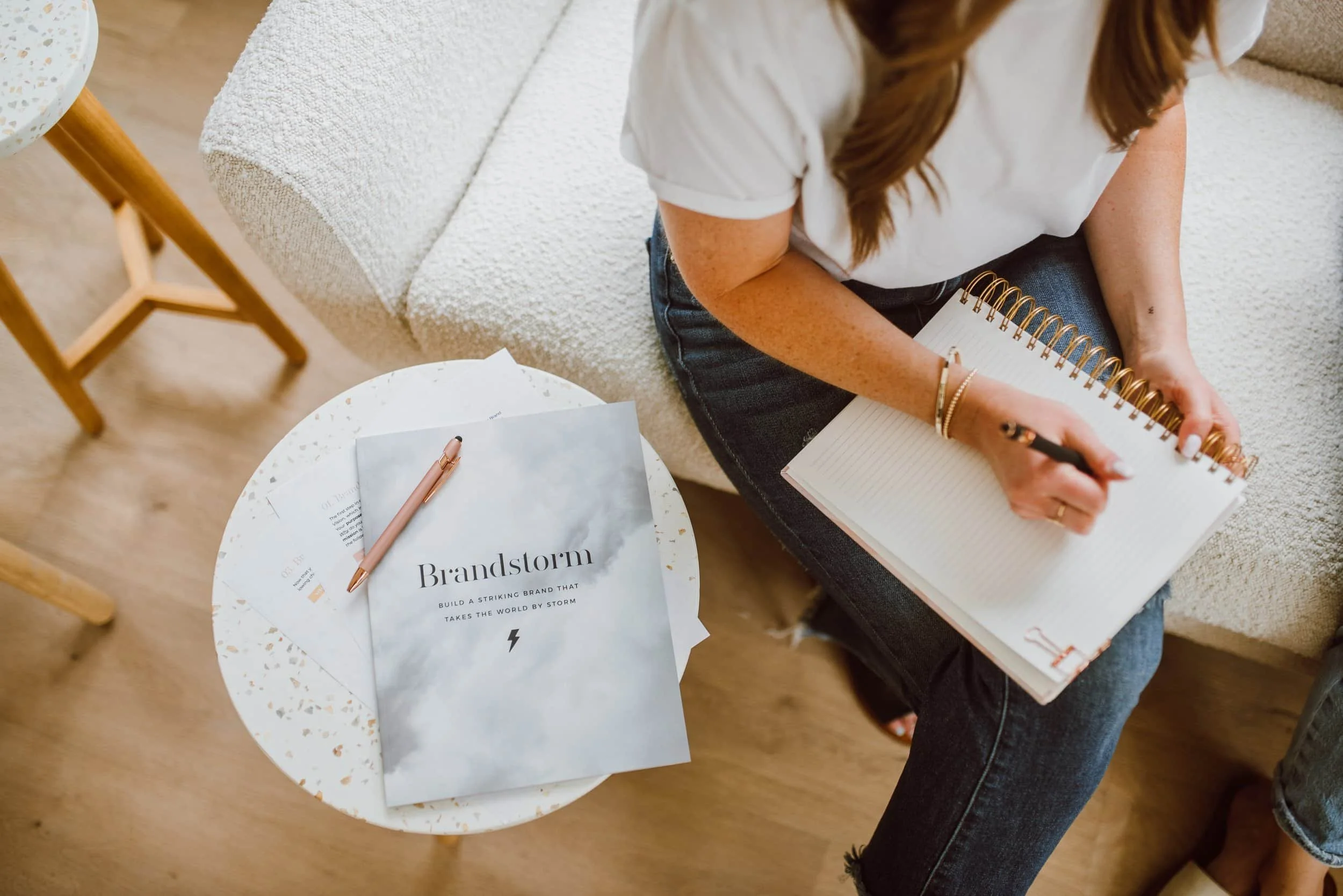 A woman with long brown hair sitting on a beige sofa, writing in a spiral notebook. She is wearing a white t-shirt and dark jeans. On a white, speckled round table beside her, there is a magazine titled 'Brandstorm' and a pink pen.