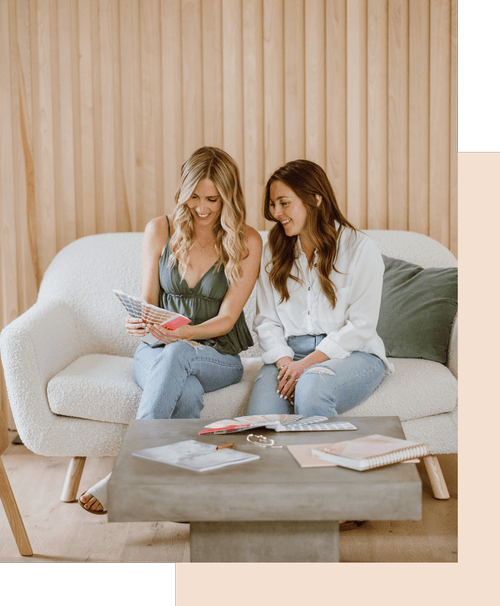 Two women sitting on a white sofa, looking at brochures, with a coffee table in front and wooden wall behind.