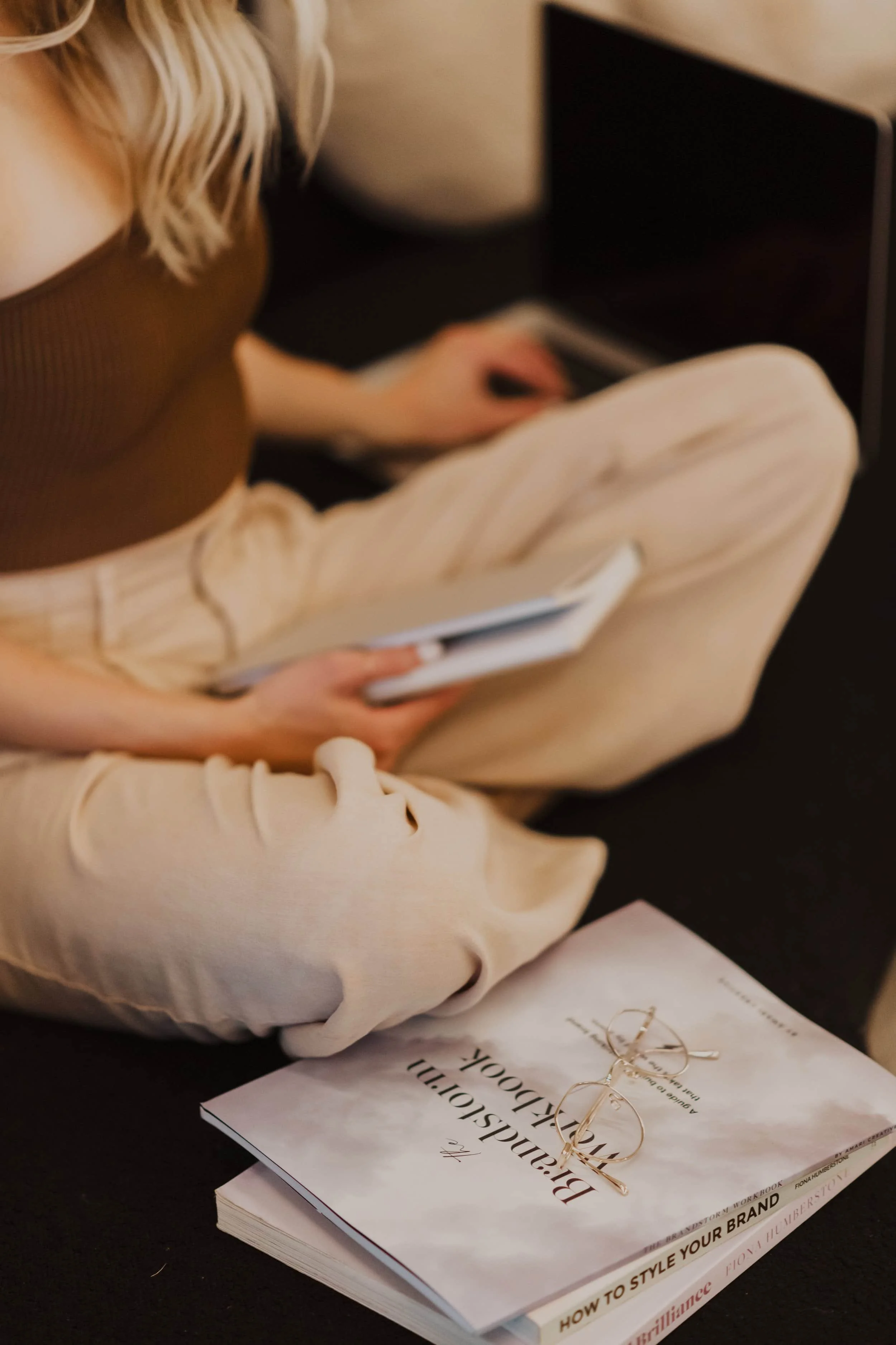A person sitting at a table with a fashion book titled 'Bridespo...' and a pair of gold earrings on top. The person is holding a phone, wearing a brown top and beige pants, with long blonde wavy hair.