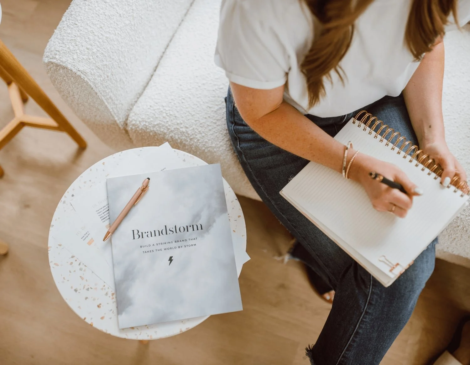 A woman sitting on a beige sofa writing in a spiral notebook with a black pen. On a white round table next to her, there is a pink pen and a stack of papers or brochures titled 'Brandstorm' with a subtitle about building a striking brand.