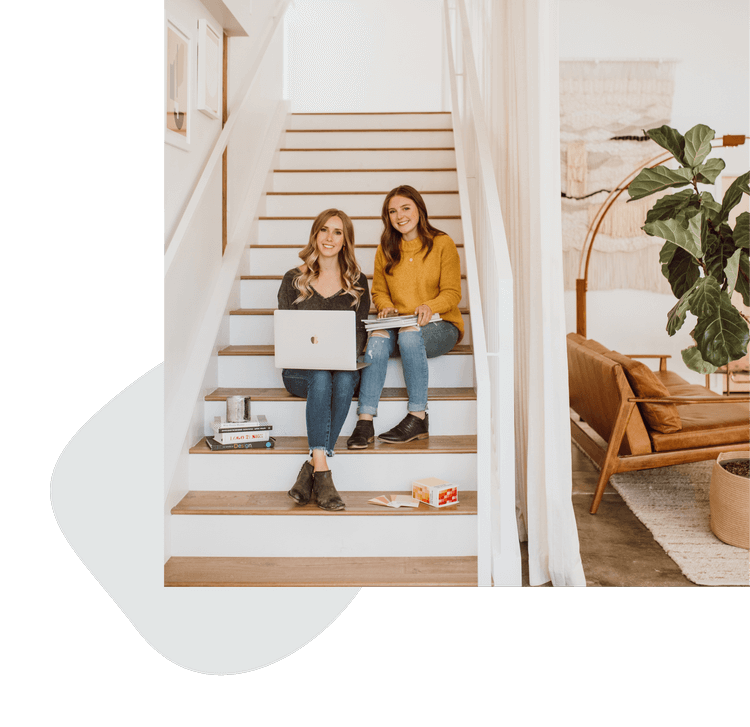 Two young women sitting on indoor staircase, smiling, with laptop and books, in a modern home with houseplants and cozy furniture.