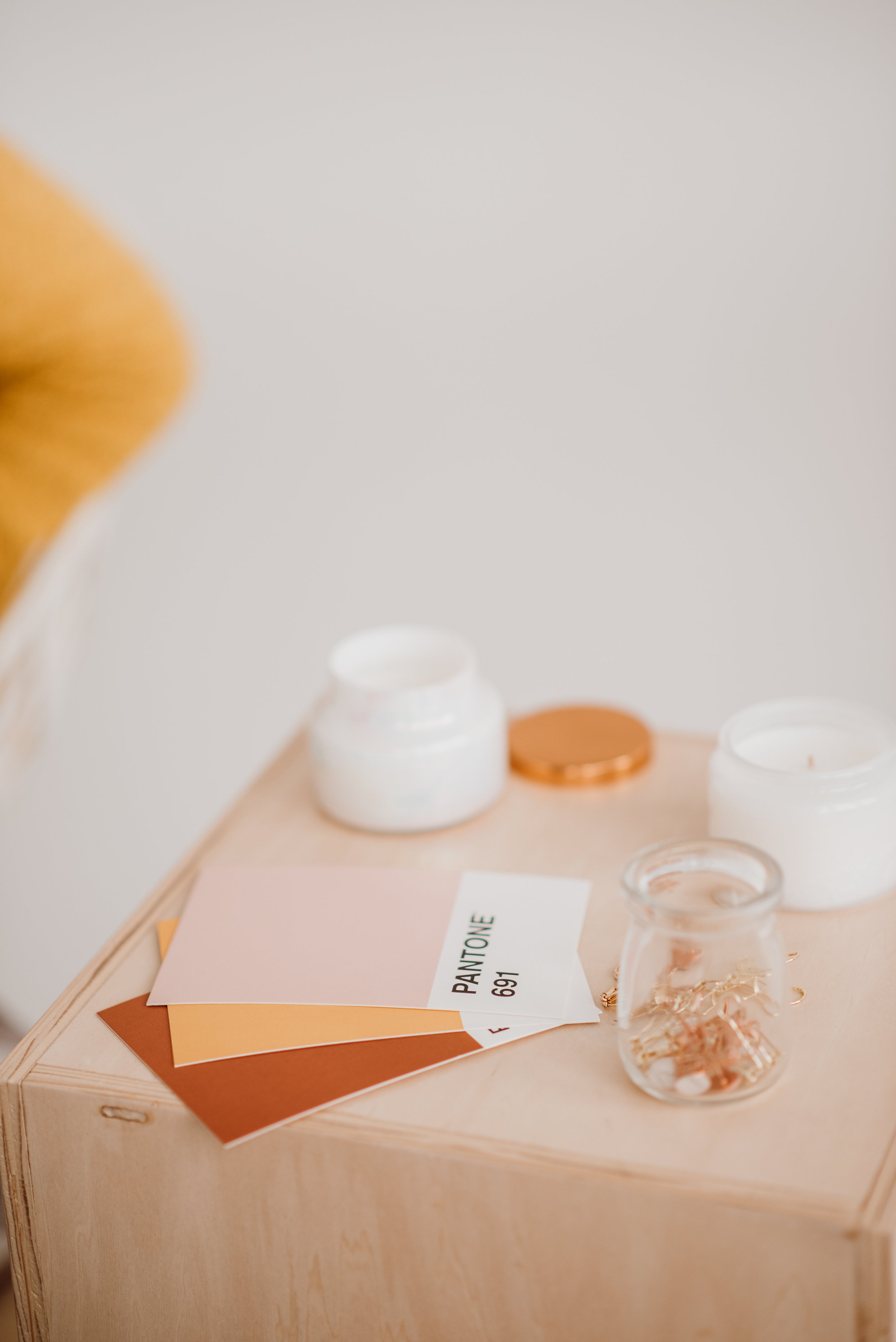 A display setup with color swatches, paint sample cards labeled 'Paintstone 691,' small containers with skincare or cosmetic products, and a clear glass beaker holding some jewelry or small metallic objects on a light wooden round table.
