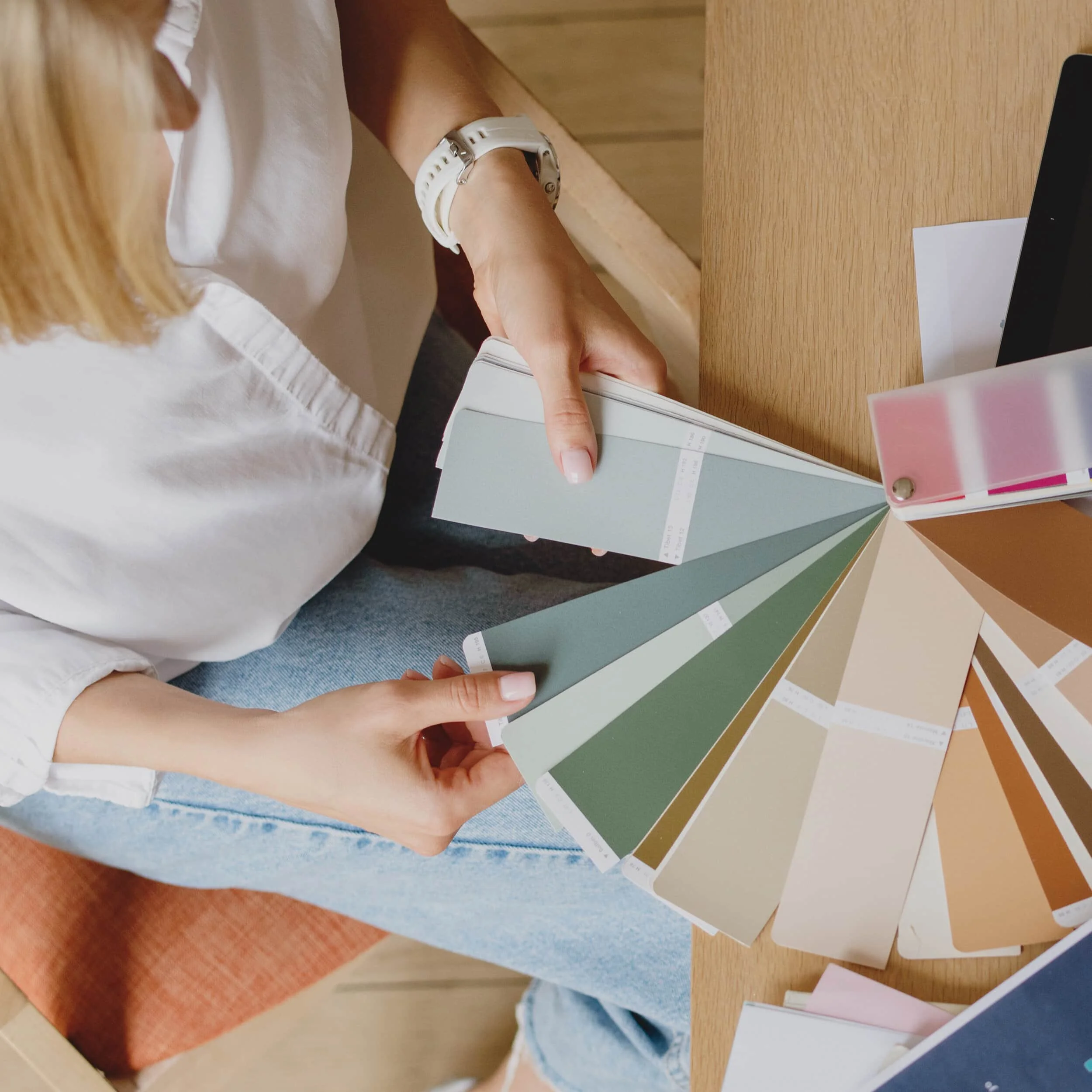 A person wearing a white watch and white shirt is holding a color swatch or paint sample card over a wooden table, with multiple other color swatches fanned out on the table.