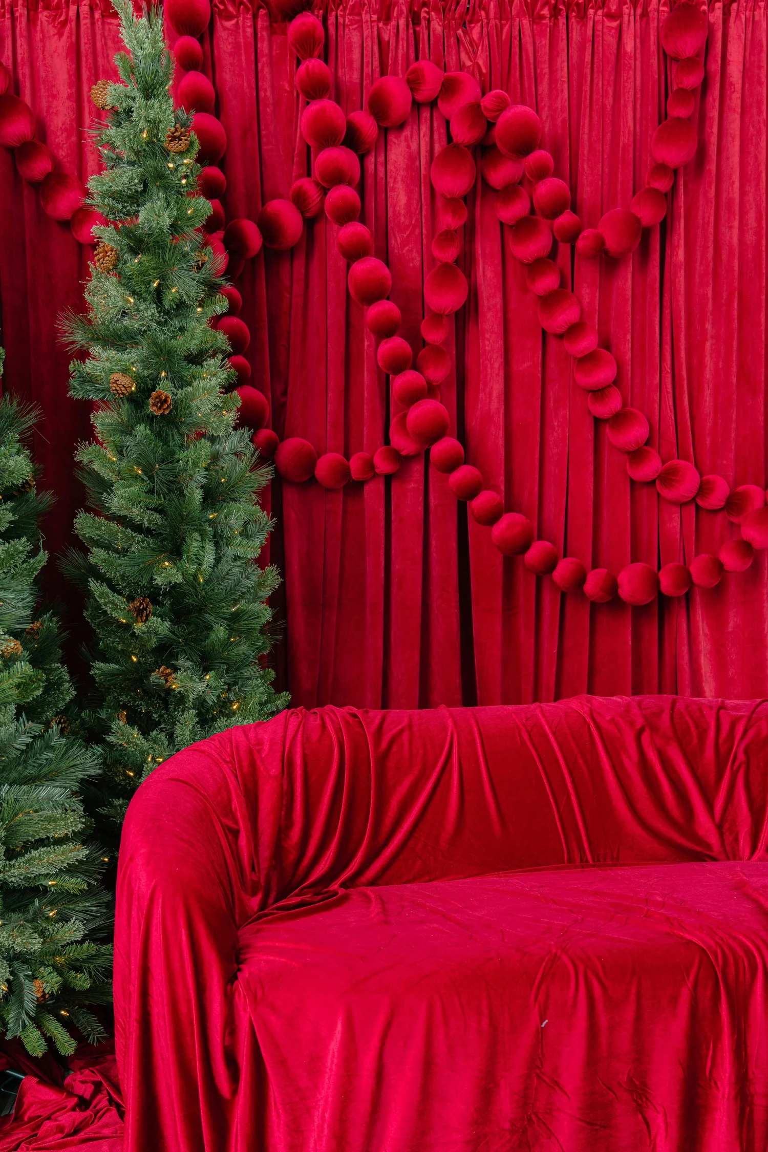 Cranberry velvet loveseat beside a lit evergreen under pom-pom garland on the Red Velvet holiday photo set in Pflugerville