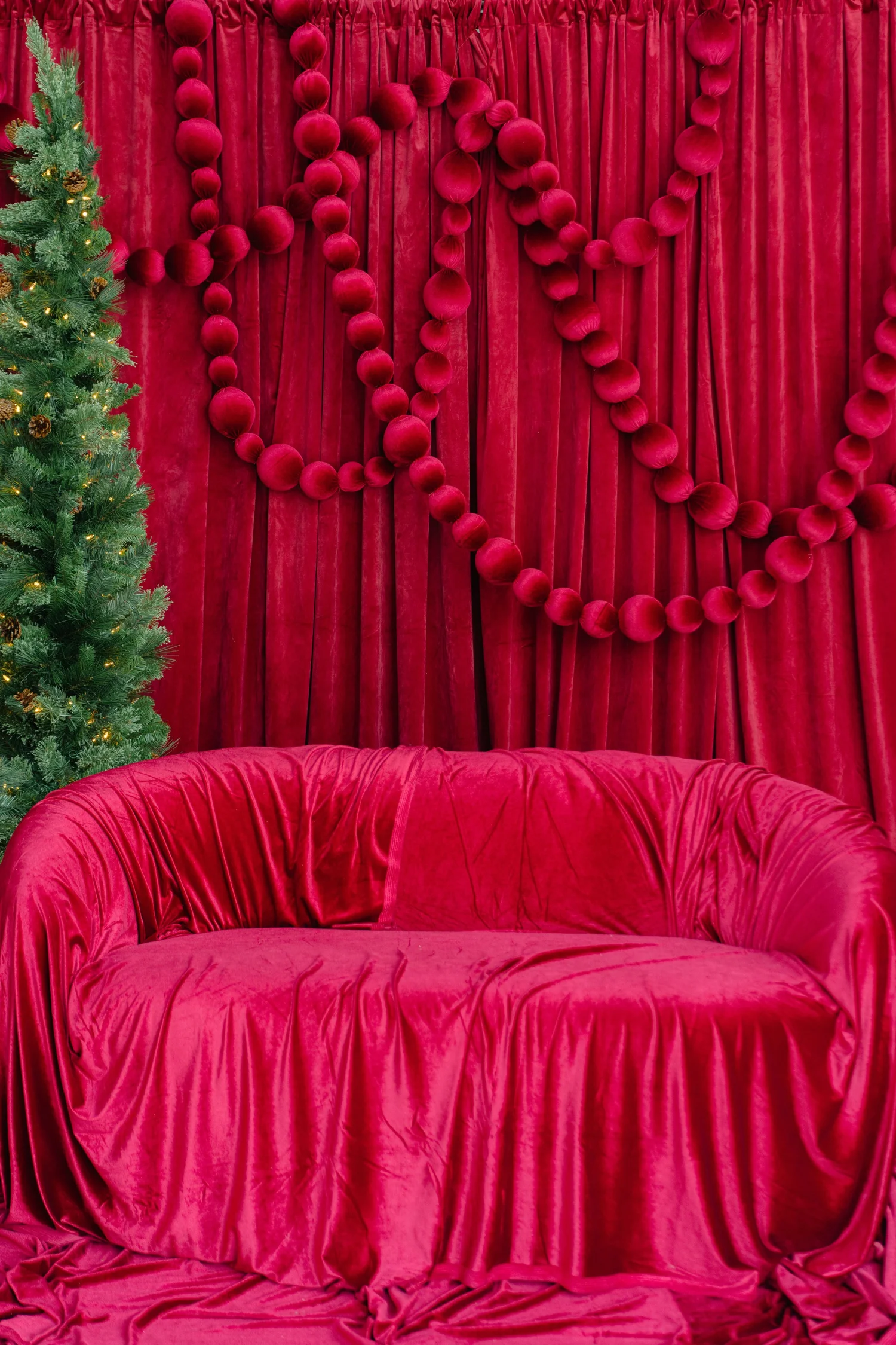 Red Velvet holiday photo set with a ruched velvet loveseat, pom-pom garland drape and lit Christmas tree at Alight Collaborative studio in Pflugerville, Austin TX