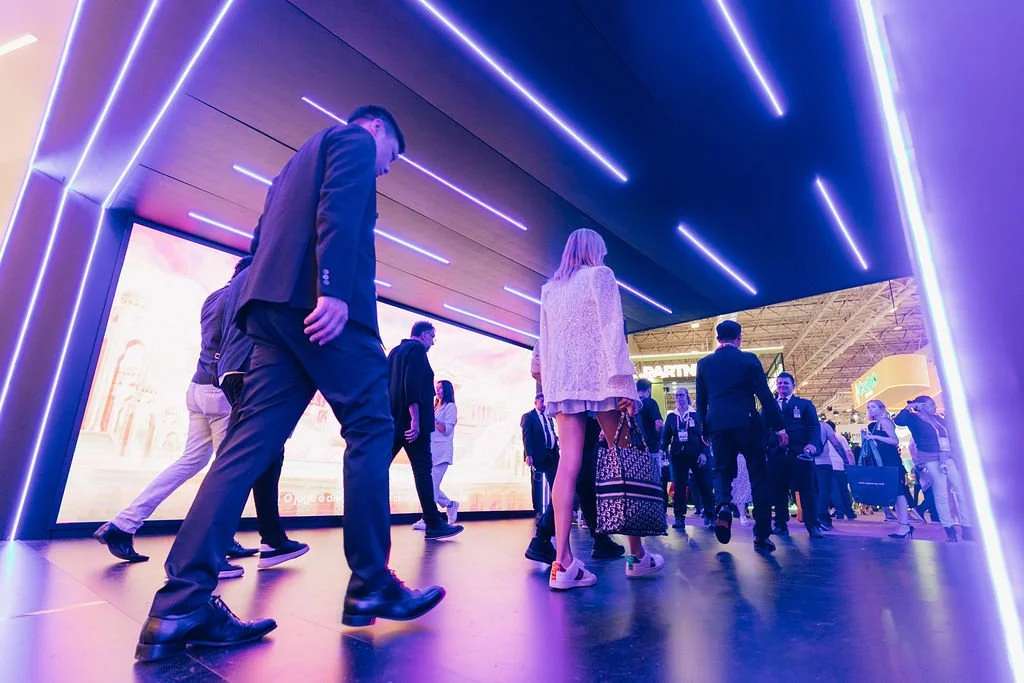 Pessoas andando em um corredor com iluminação neon azul e uma grande tela de vídeo ao fundo.