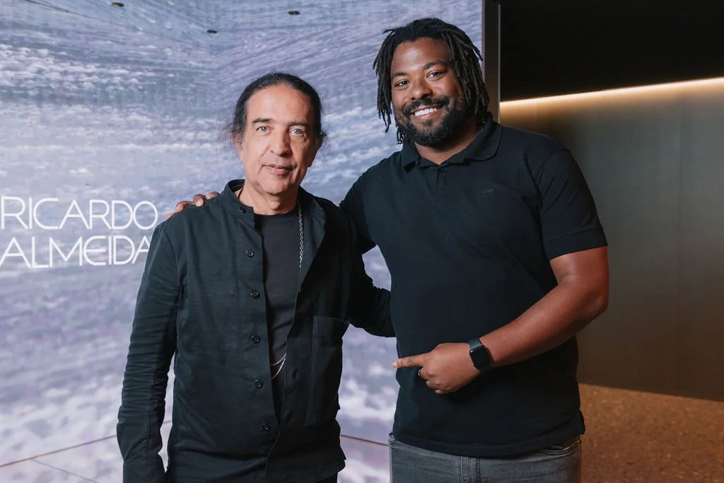 Duas pessoas, um homem mais velho e um mais jovem, sorrindo, posando juntos para uma foto, com fundo digital exibindo parcialmente o nome 'Ricardo Almeida'.