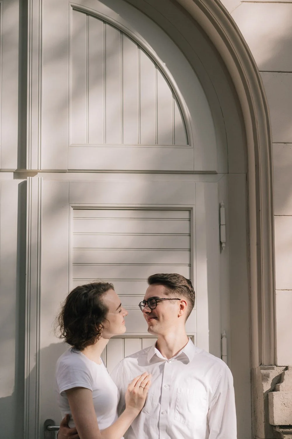 Casal sorridente se olhando em frente a uma porta branca com detalhes clássicos.