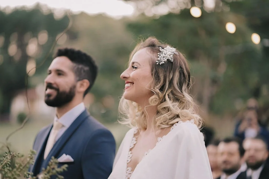 Noivos sorrindo em uma cerimônia ao ar livre, com o homem de barba e terno azul escuro e a mulher de cabelo cacheado usando um vestido branco e acessório na cabeça, ao fundo com pessoas assistindo.